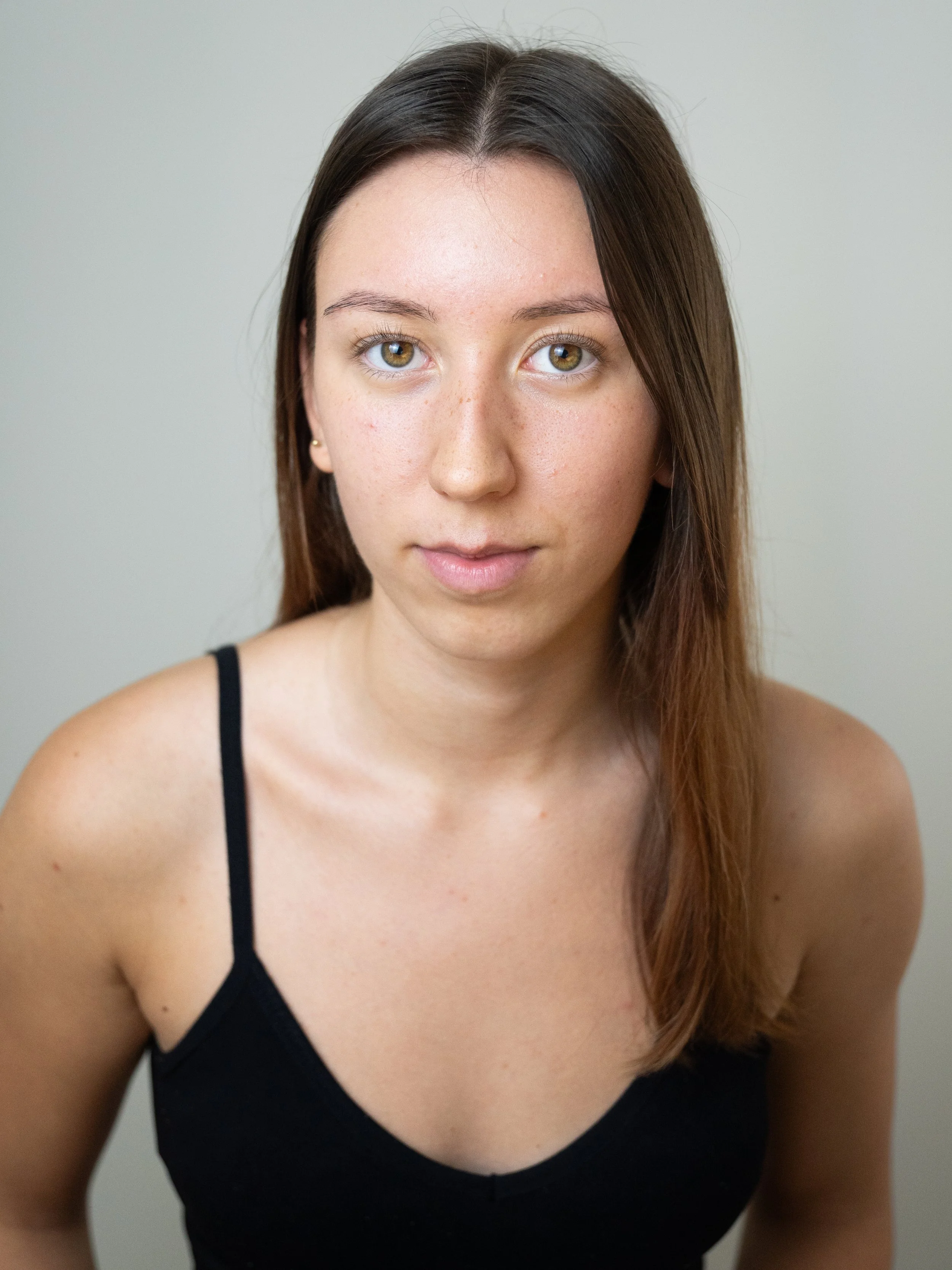 A young woman with long brown hair, light skin, and hazel eyes wearing a black spaghetti strap top, standing against a plain light-colored background.