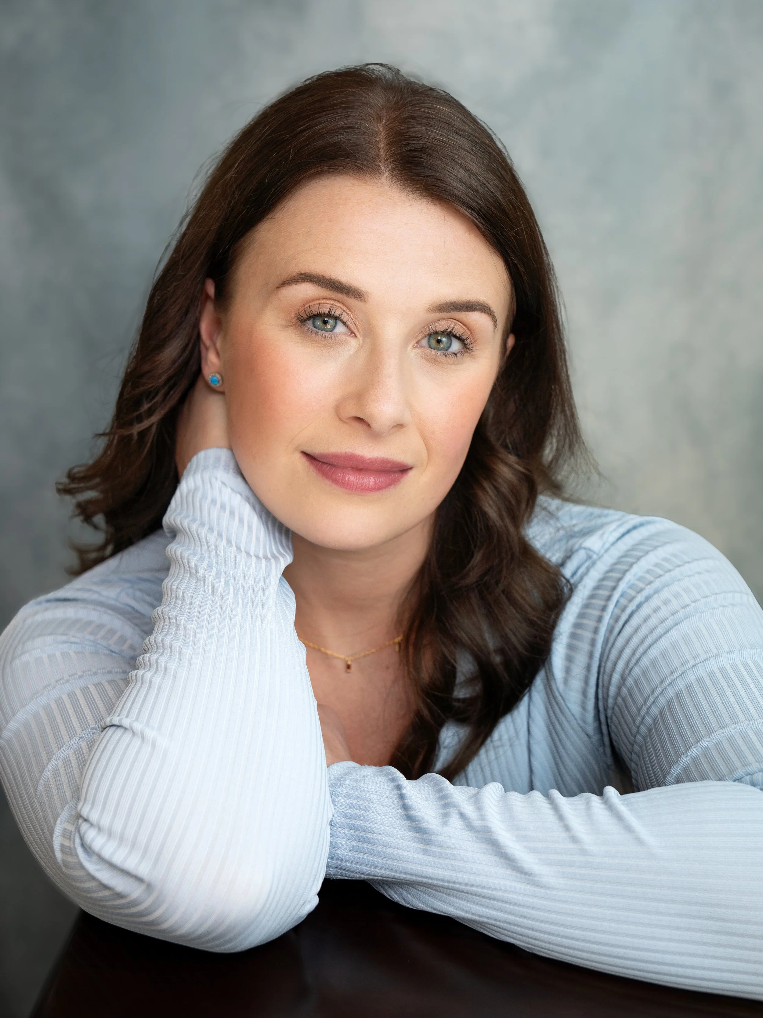 An actors heashot of a woman with dark brown hair. She is in a lightblue top, sitting infront of a light bllue background. She is leaning on her right arm with a soft expression on her face. 