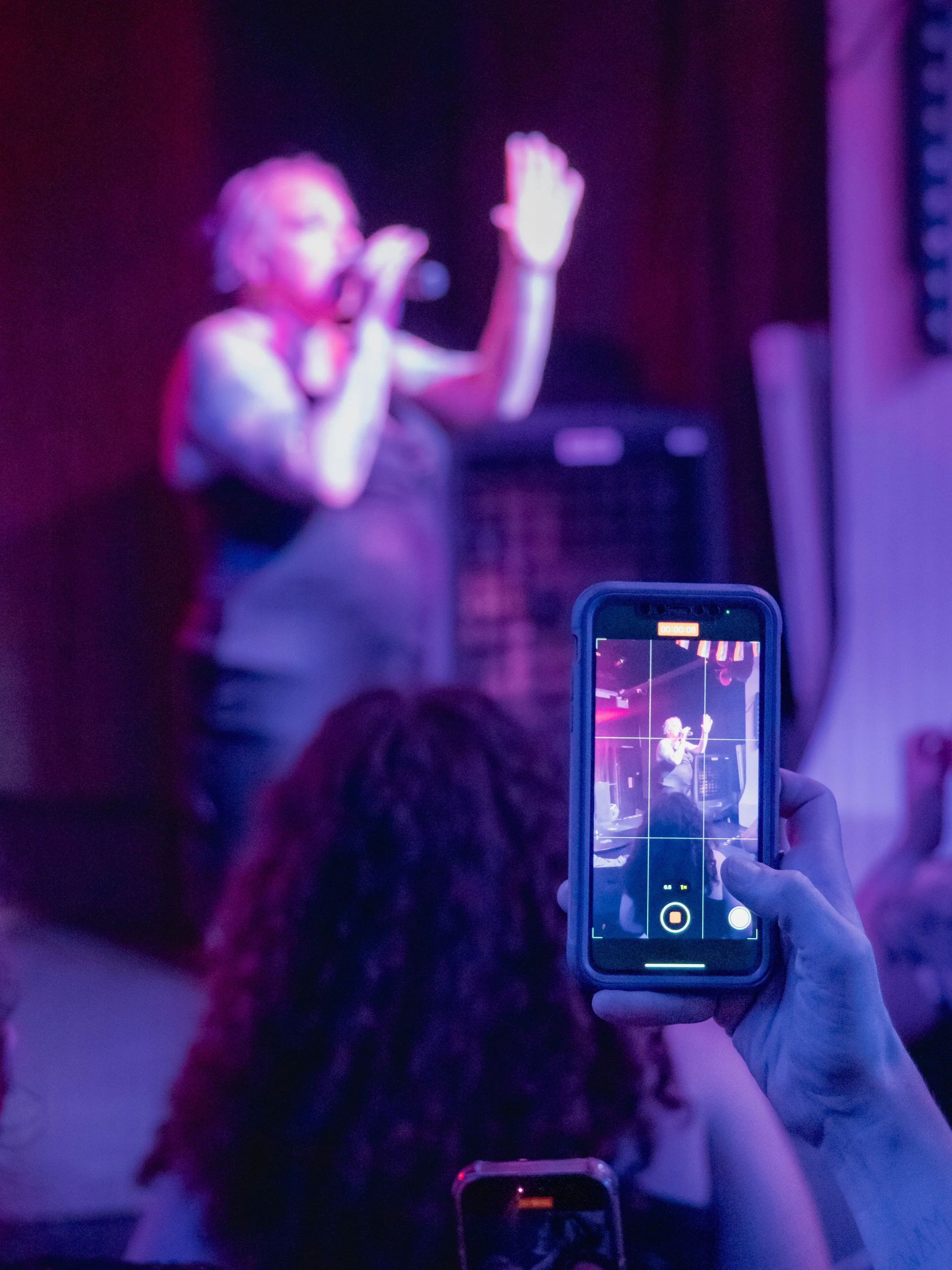 A person is recording a woman singing into a microphone on stage, with the scene displayed on a smartphone screen in a dimly lit venue.