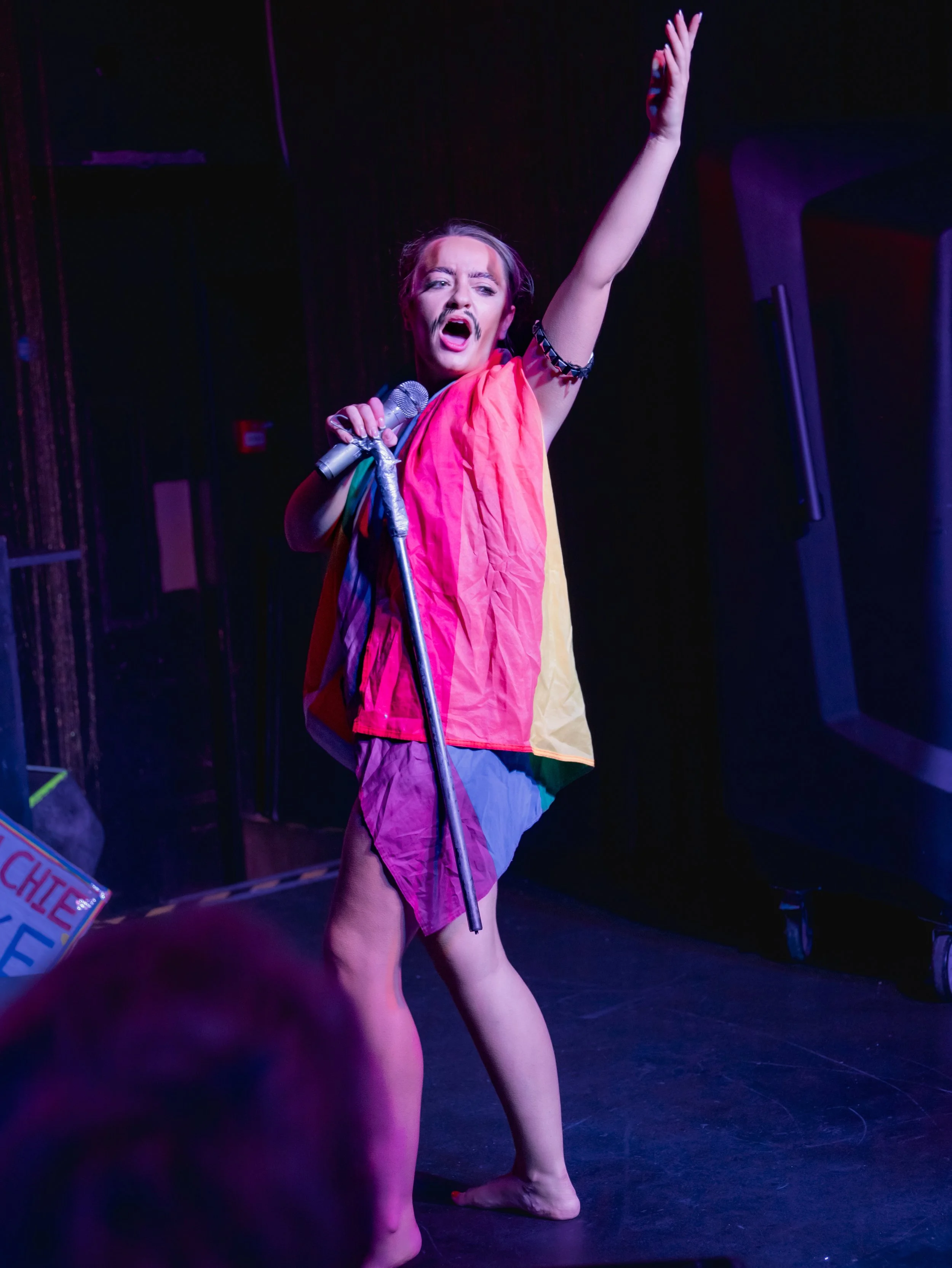 Performer on stage wearing colorful clothing, holding a microphone, with one arm raised, singing passionately.