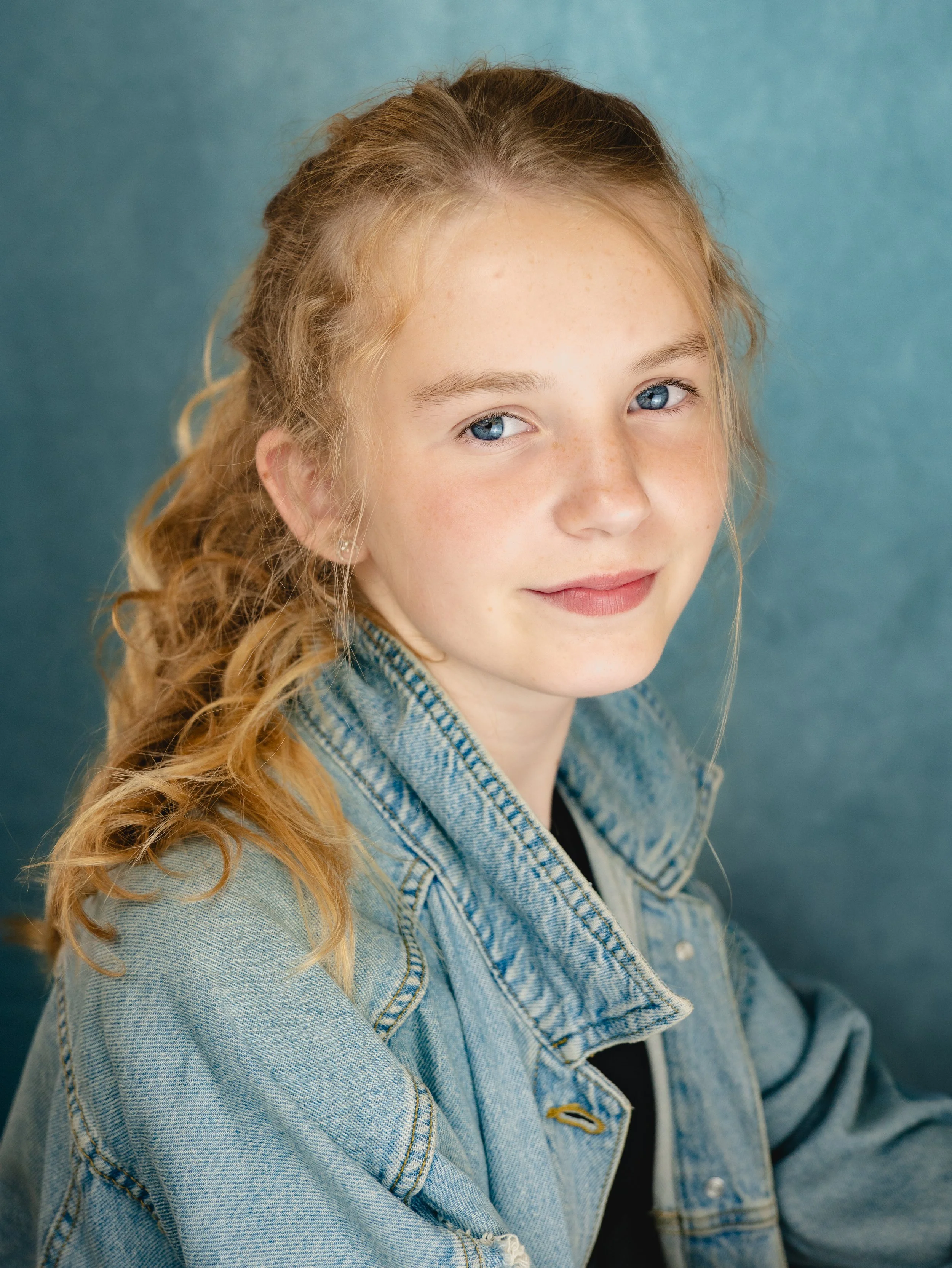 A young girl with red hair, blue eyes, and freckles, wearing a denim jacket, smiling softly, against a textured blue background.