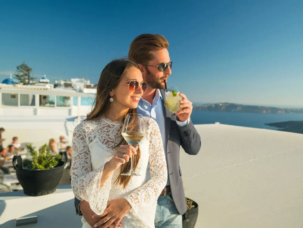 Ein junges Erwachsenes Paar trinkt Getränke auf einer Terrasse mit Meerblick bei Sonnenschein. Und geniesst das Leben. Vorsorge, Vermögensaufbau, Anlagen, Erben.