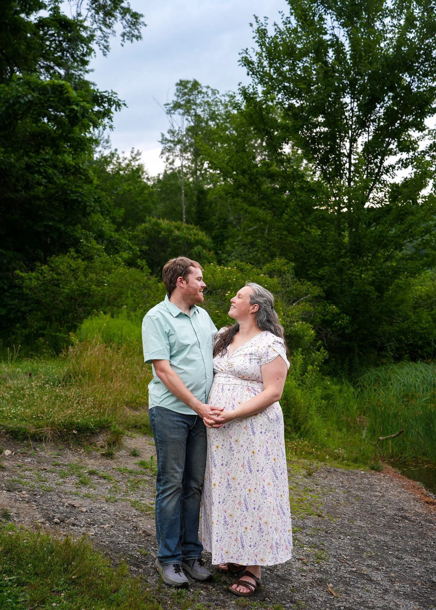 A couple holding hands and looking at each other, standing outdoors in a natural, green setting with trees and grass around them.