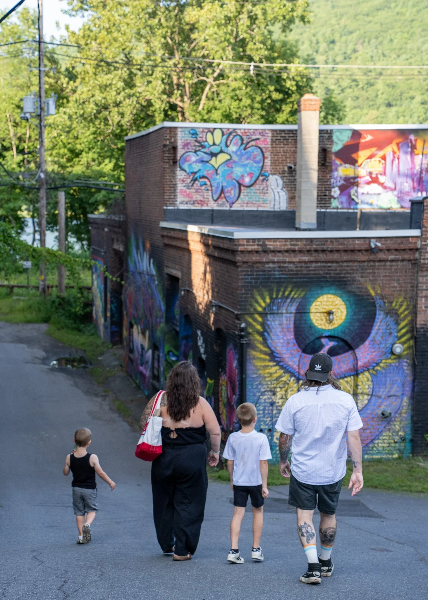A group of people walking on a paved street past a building with colorful murals. The group includes adults and children, some with tattoos and distinctive clothing. The area is green and leafy, with trees in the background.