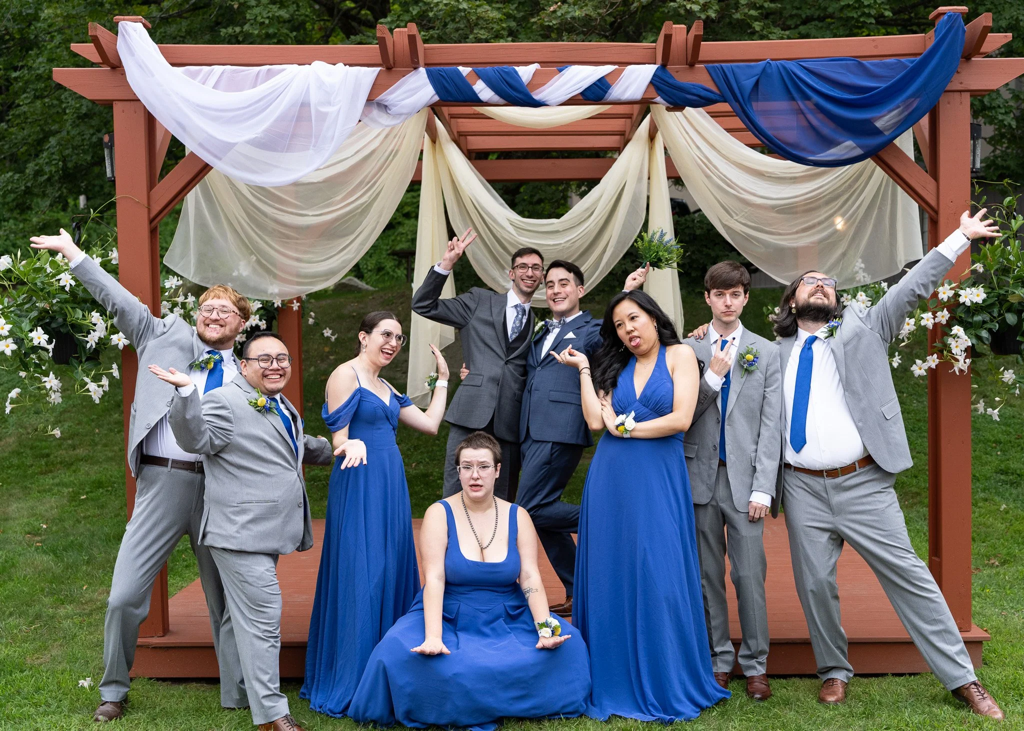 A group of people dressed in blue and gray formal attire posing under a decorated wooden archway with curtains and greenery in the background.