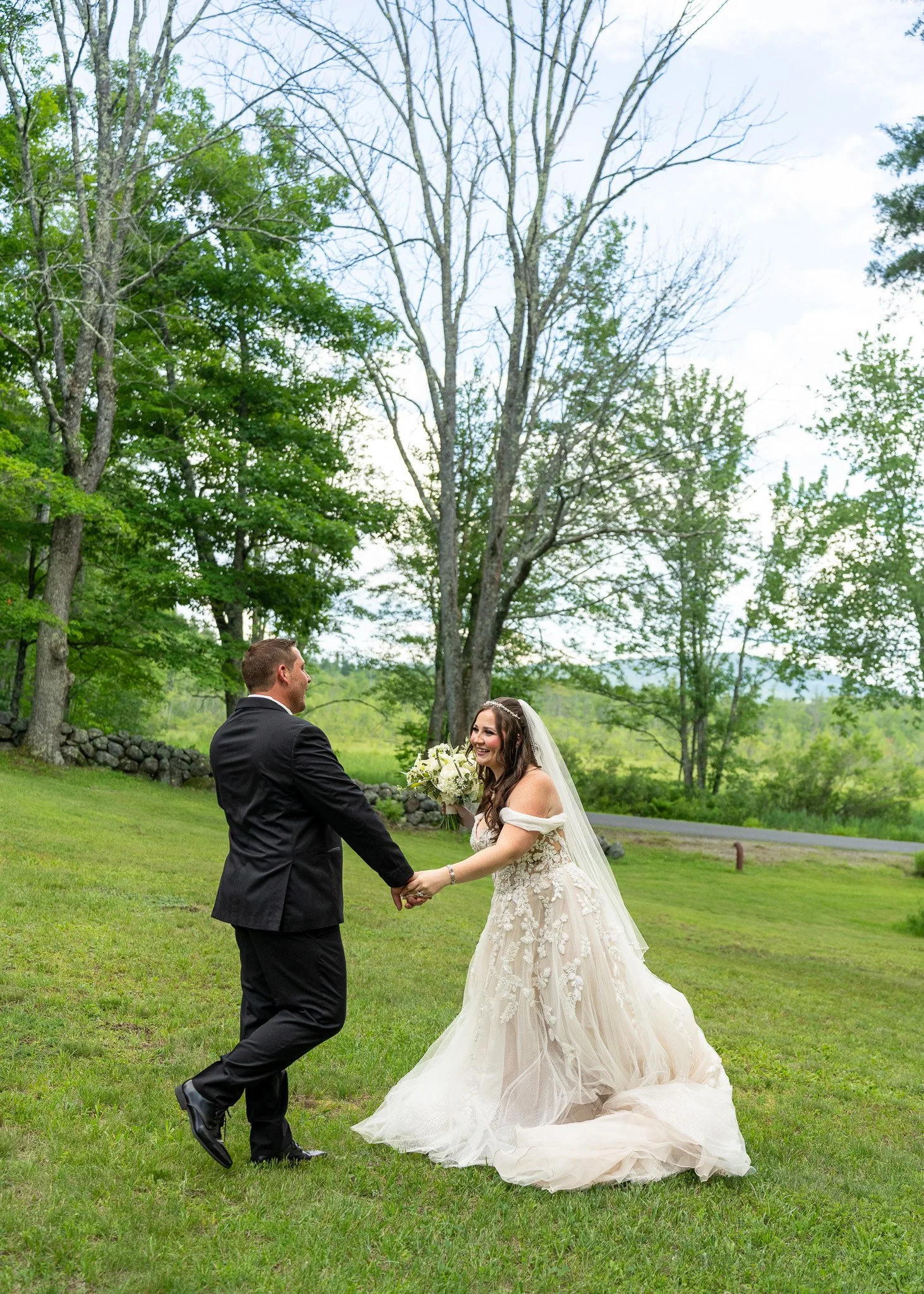 A bride and groom holding hands outdoors in a grassy area during daytime, with trees and a stone wall in the background. The bride is wearing a white wedding gown and veil, and the groom is in a black suit. They are smiling and looking at each other.