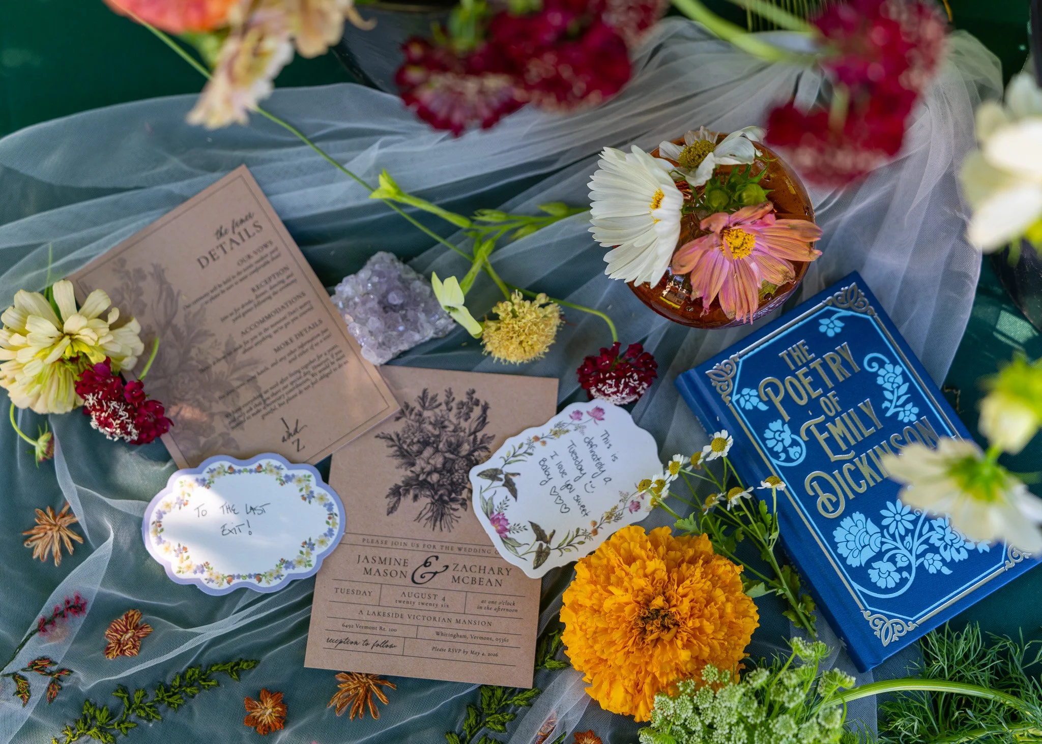 A wedding table display with flowers, wedding invitations, a book titled 'The Poetry of Family and Happiness,' and decorative papers. The flowers include daisies, marigolds, and various colorful blooms.