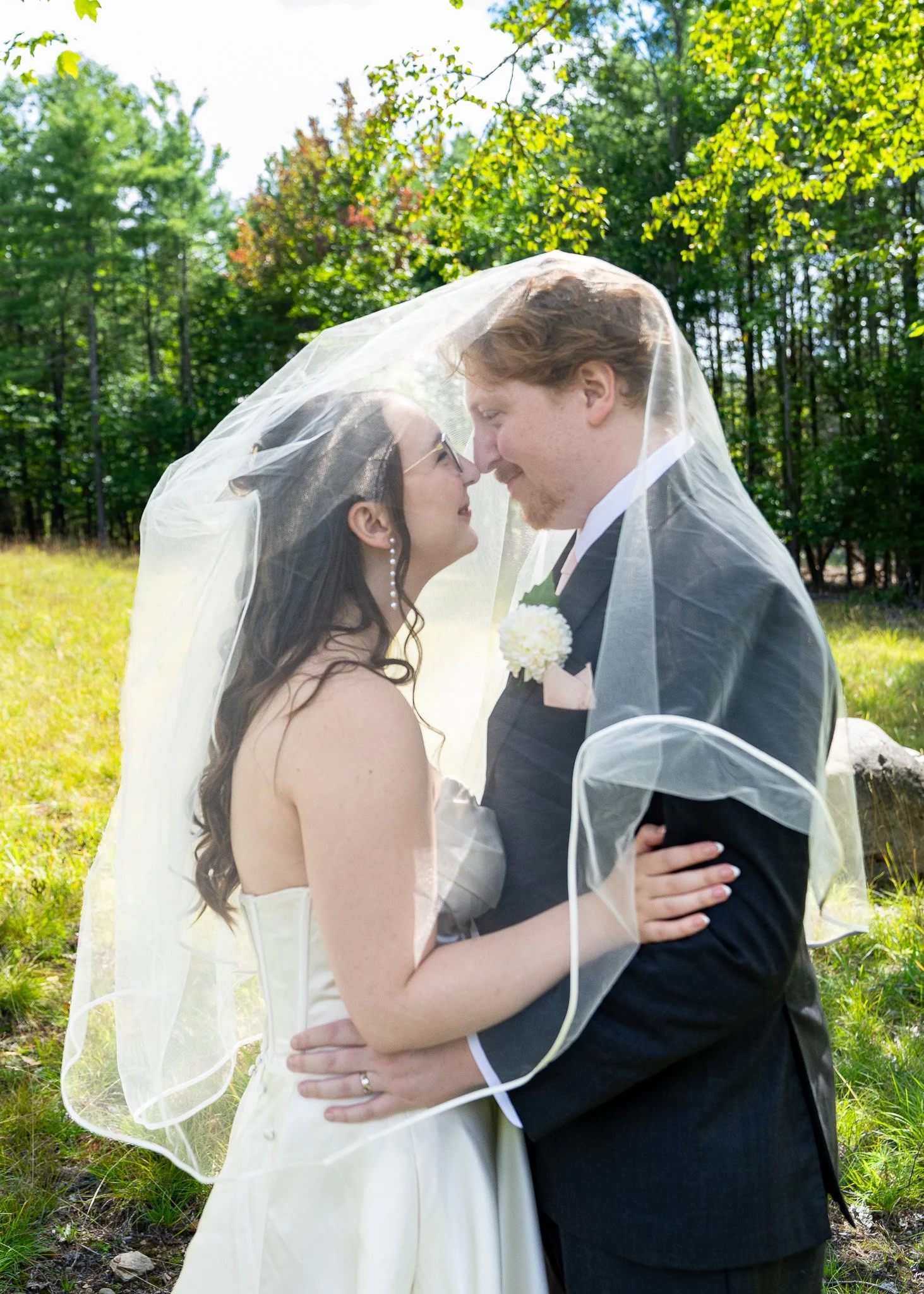 A bride and groom standing close together outdoors under trees, sharing a tender moment with foreheads touching and smiling, under the brides sheer veil.