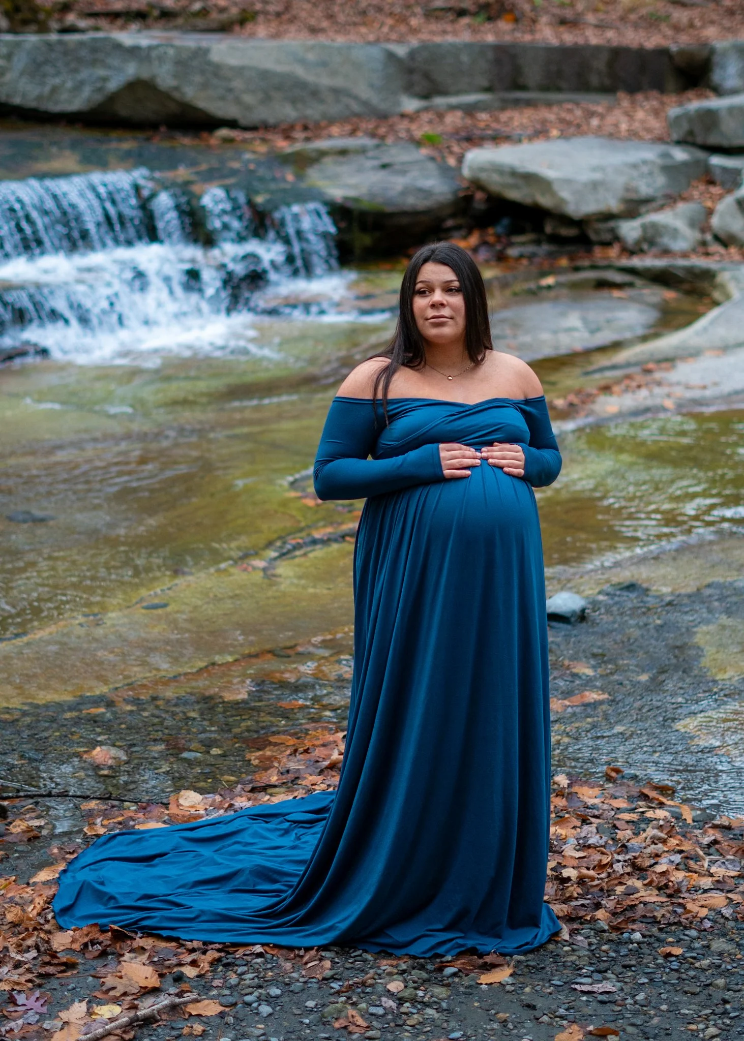 A pregnant woman in a long, off-the-shoulder blue dress standing near a small waterfall in a natural setting surrounded by fallen autumn leaves, taken at one of my favorite local Southern Vermont spots, Old Jelly Mill Falls.