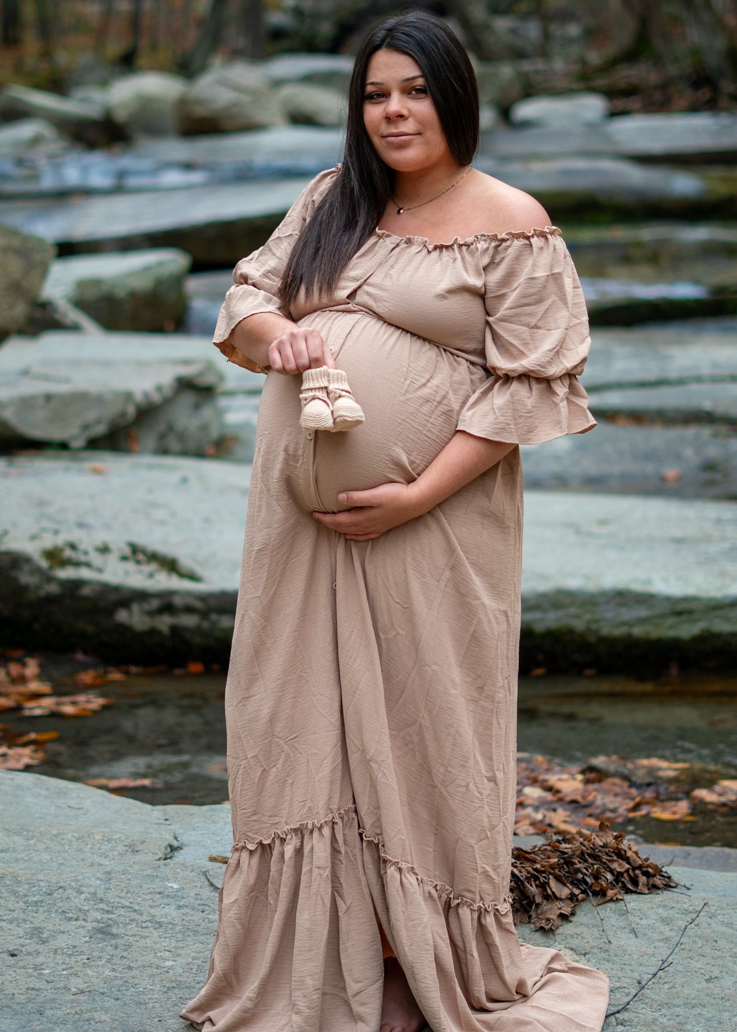 Pregnant woman standing on flat rocks near a stream, holding a pair of baby booties and touching her belly. Taken in Southern Vermont at Old Jelly Mill Falls.