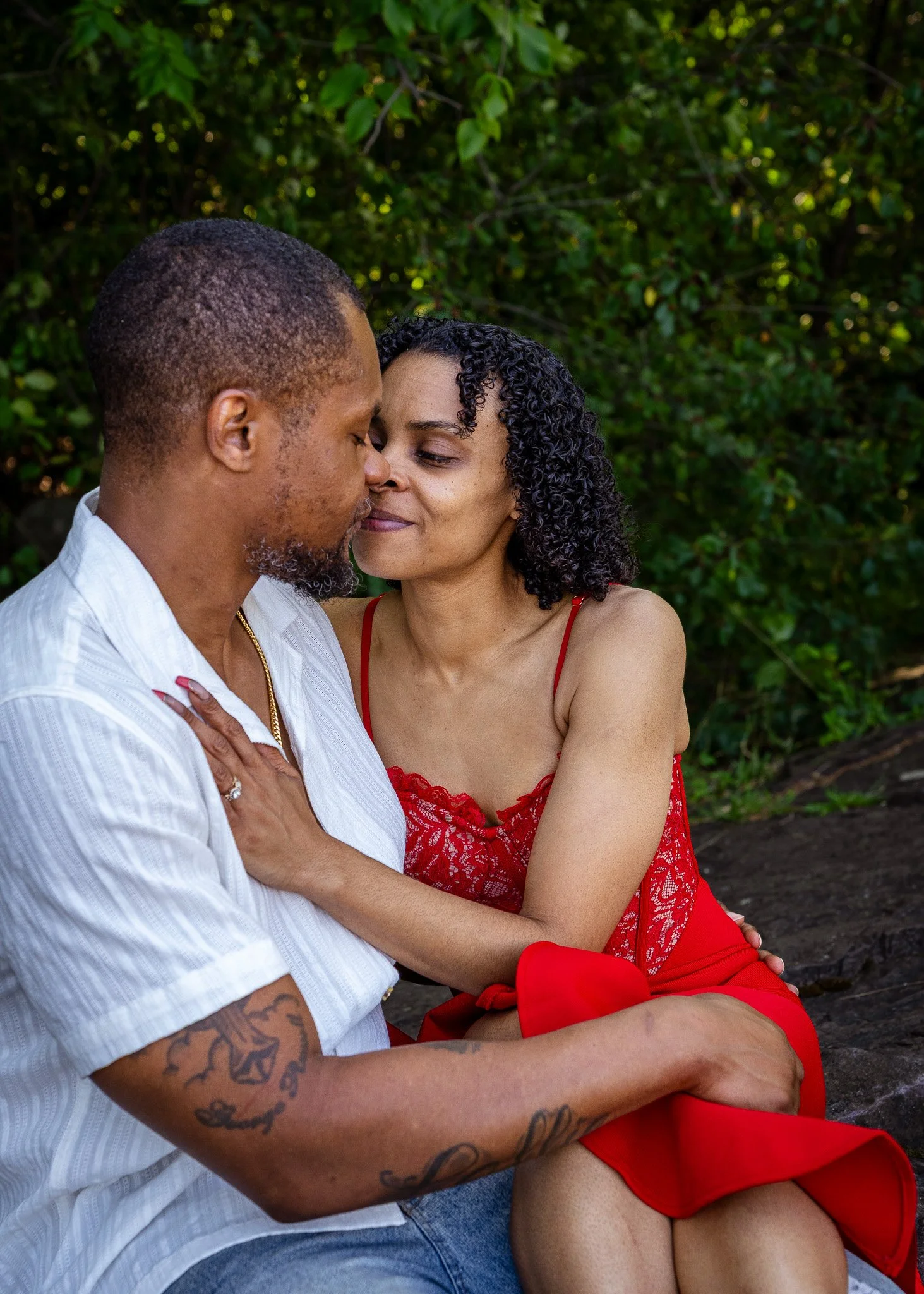 A man and woman sitting close together outdoors, touching foreheads and smiling, with green foliage in the background.