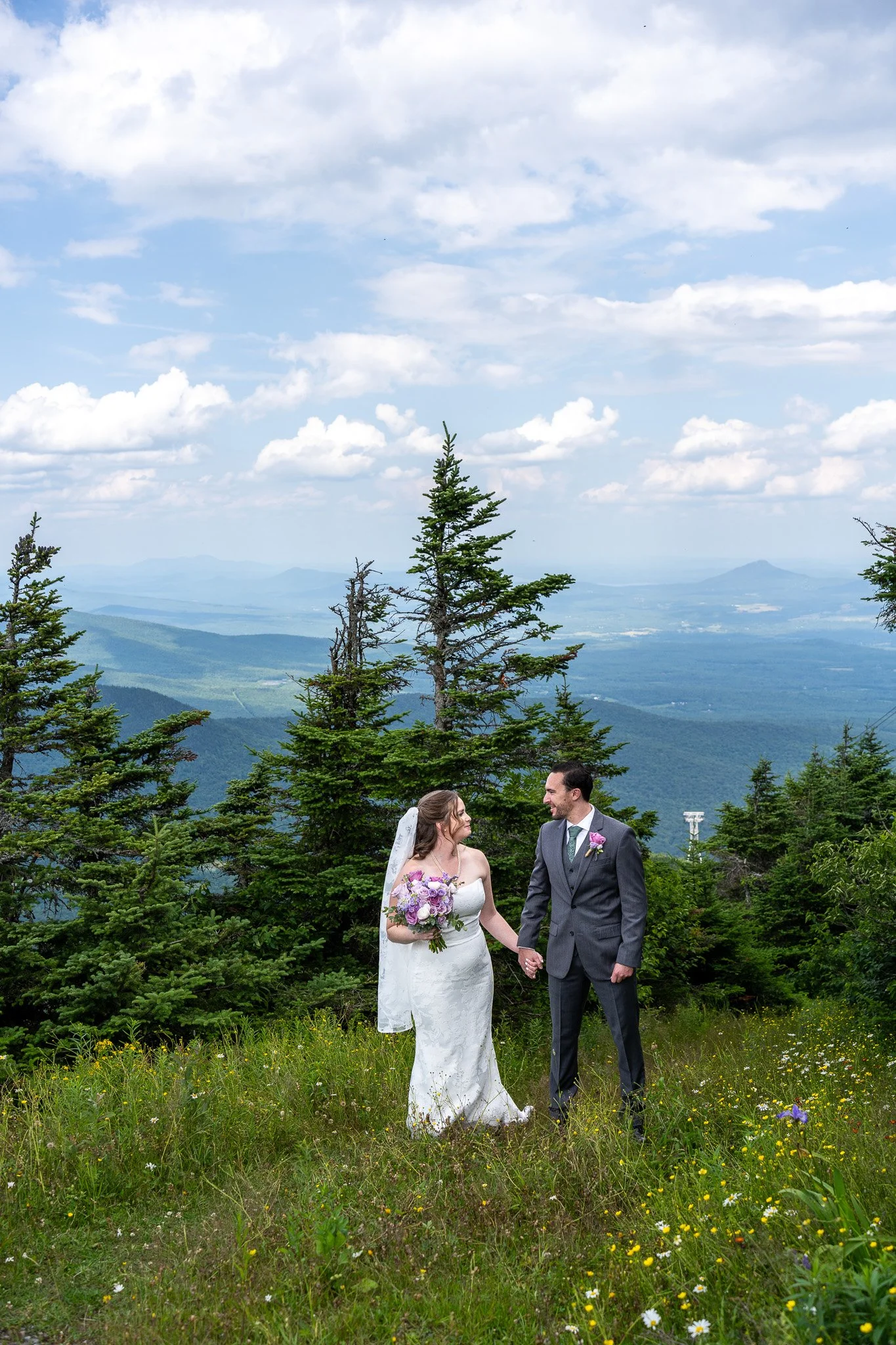 A bride and groom holding hands and smiling at each other in a lush green, mountainous landscape with trees and a cloudy sky.