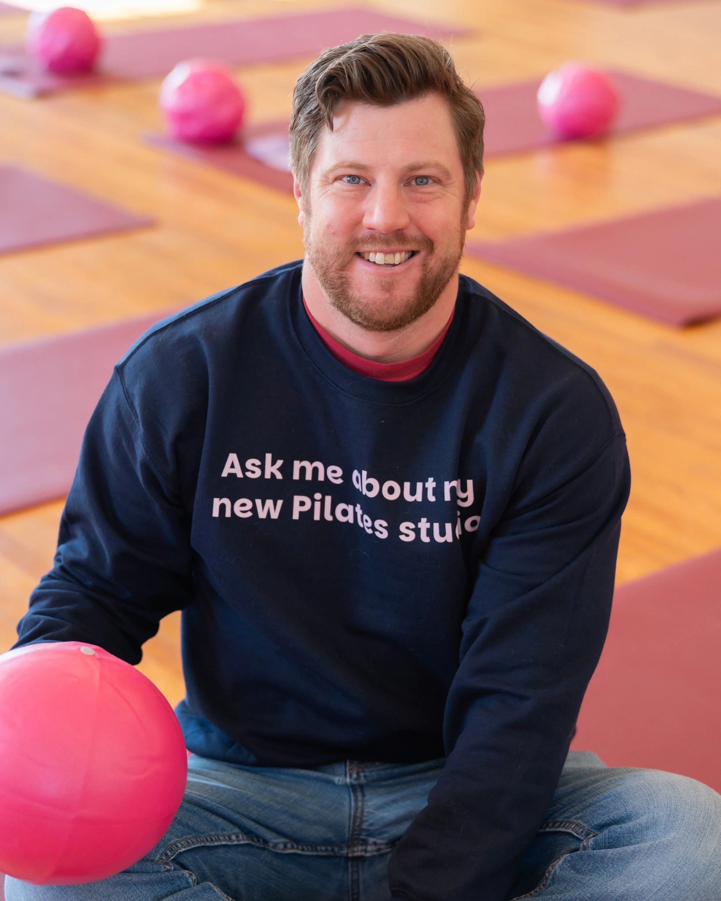 A man sitting on the floor in a Pilates studio, smiling, wearing a navy sweatshirt with white text that says 'Ask me about my new Pilates studio', with pink exercise balls and yoga mats in the background.