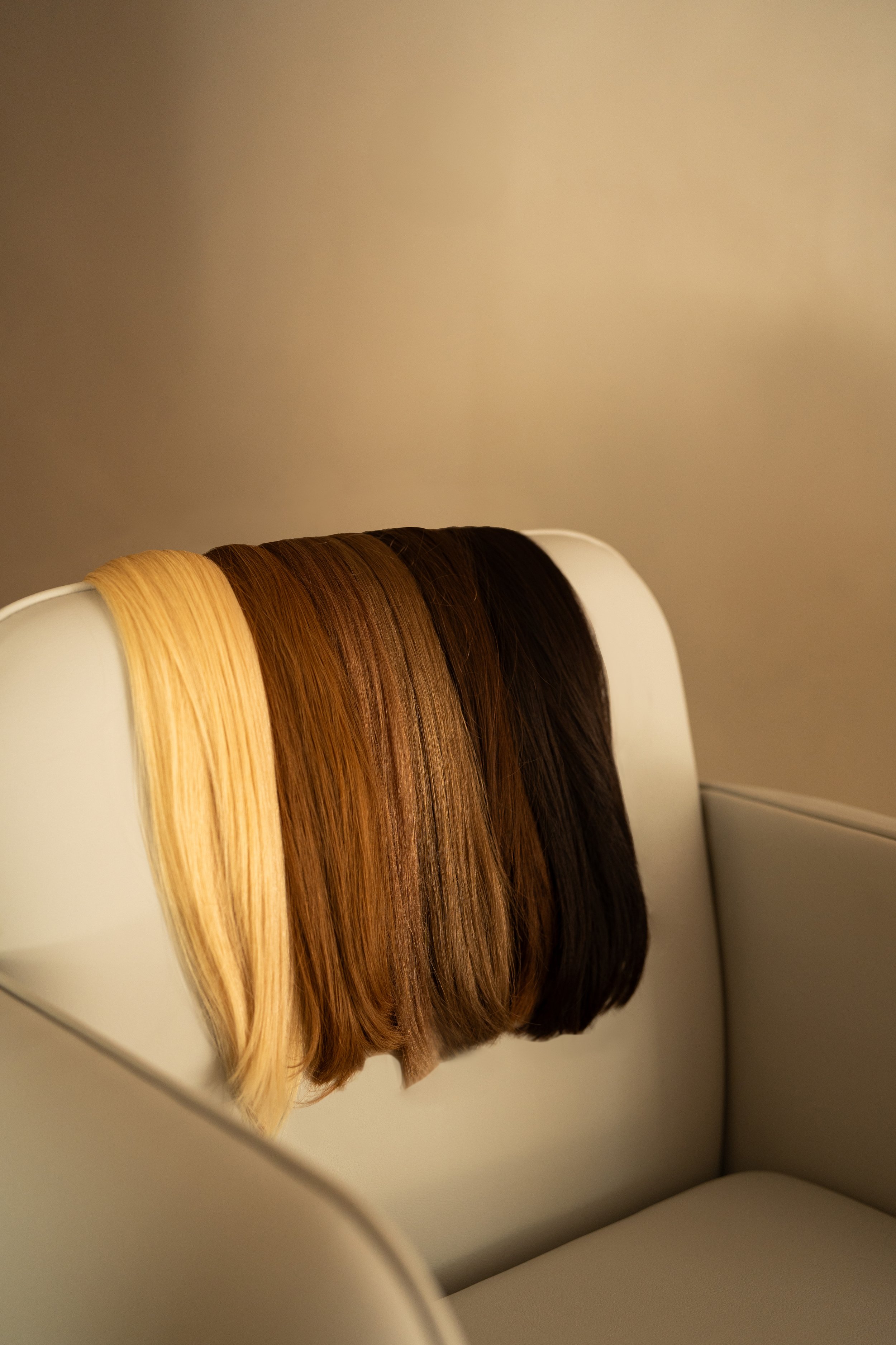 Três perucas com diferentes tons de cabelo, uma loira, uma castanha e uma negra, colocadas sobre uma cadeira de couro branca.