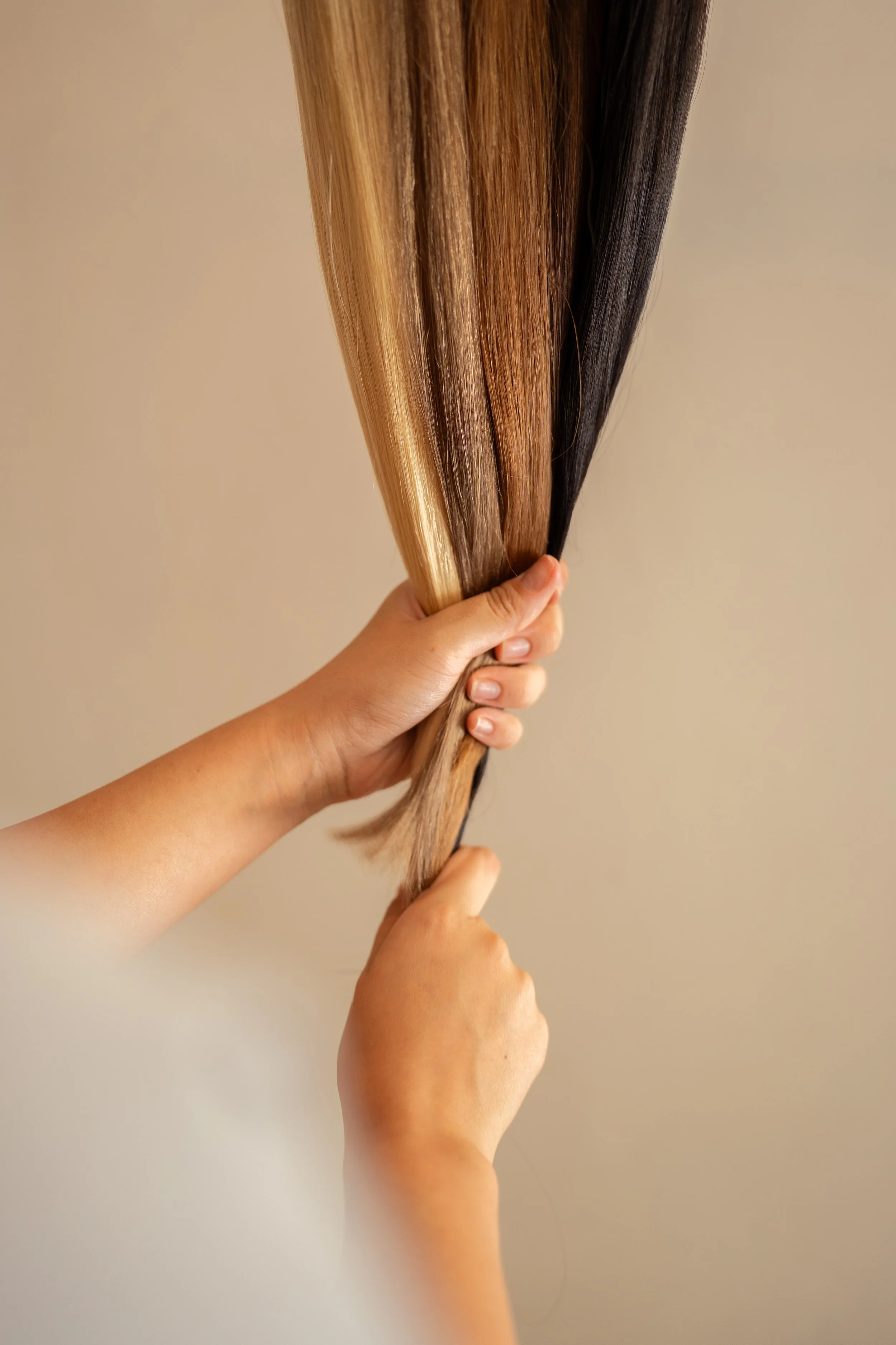 Duas mãos segurando fios de cabelo de cores diferentes, incluindo loiro, castanho e preto.