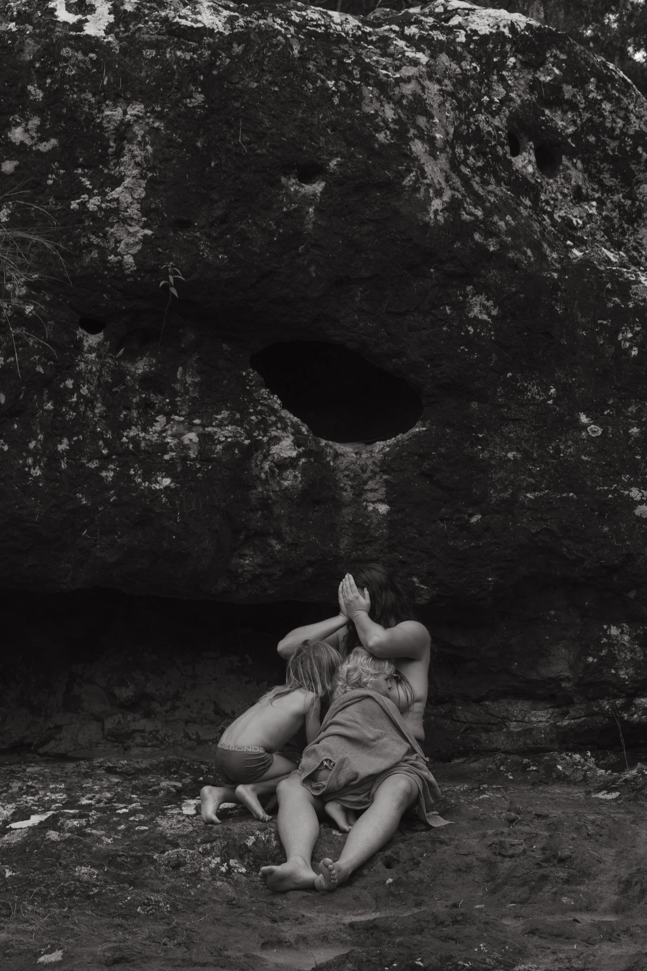Three children huddle under a large rock formation, with one covering their face with their hands, in a black and white photo.
