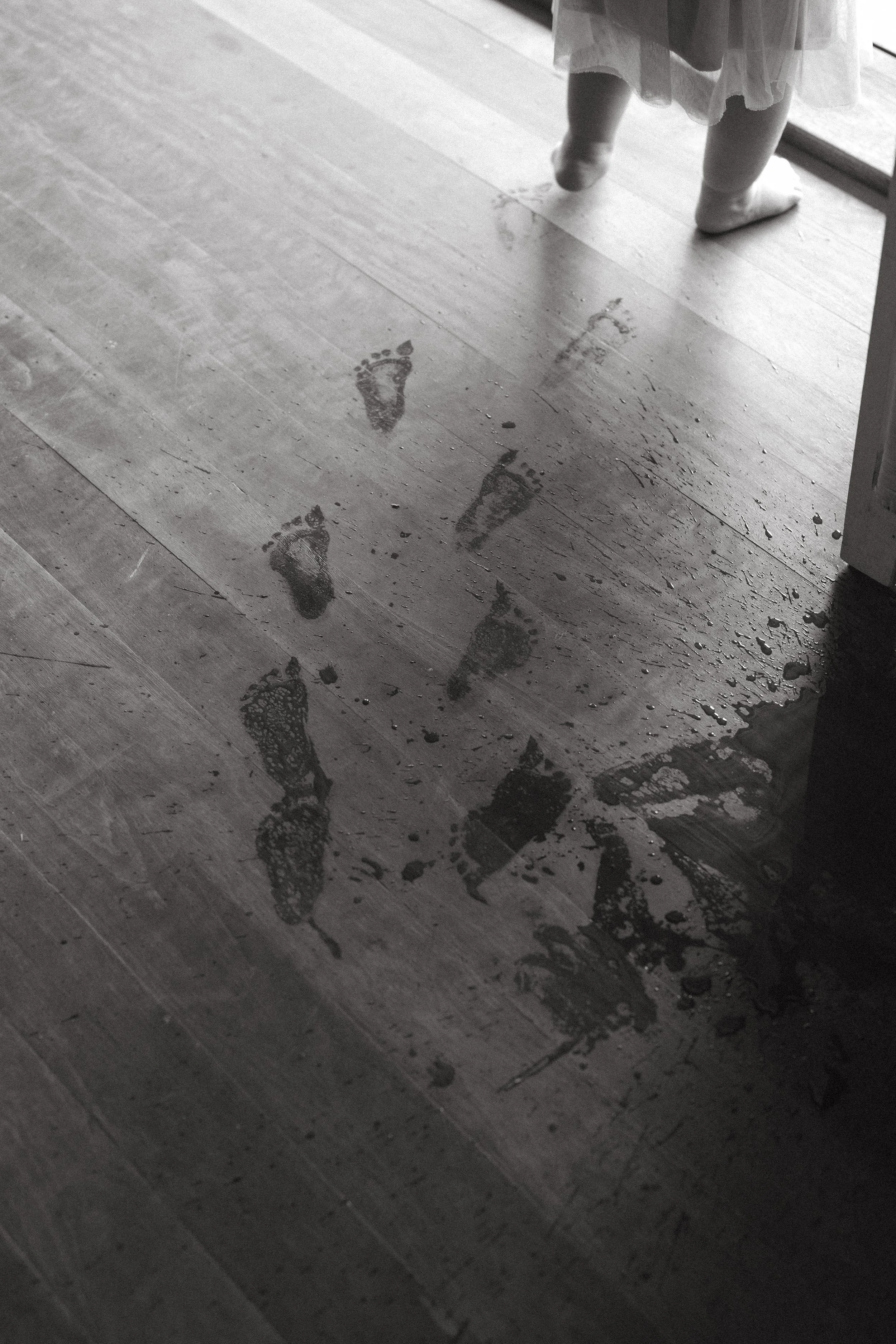 Footprints on a wet wooden floor near a child's feet wearing socks and a dress.