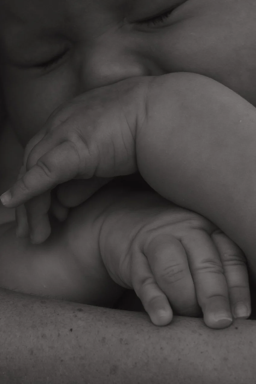 Close-up black and white photo of a person's face and hands, with one hand holding a newborn baby's hand.