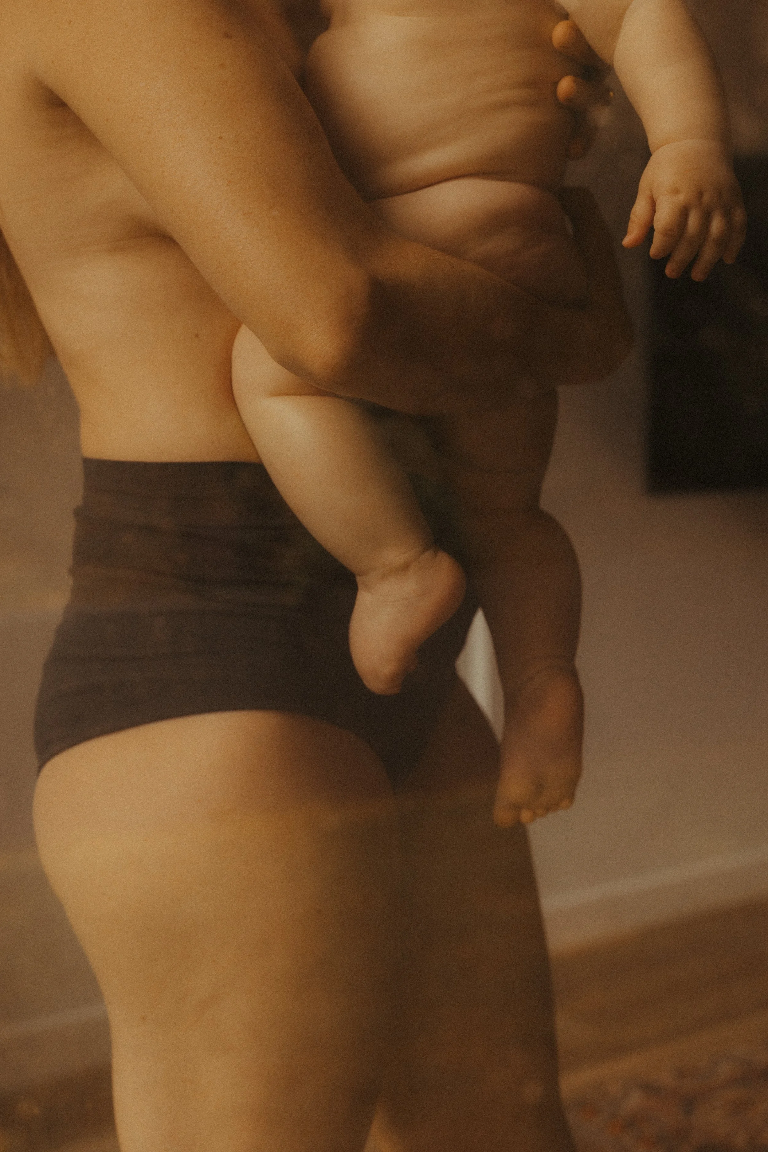 A person holding a baby, focusing on their torso, arms, and legs, with the baby’s arm and legs visible. The person is wearing black shorts.