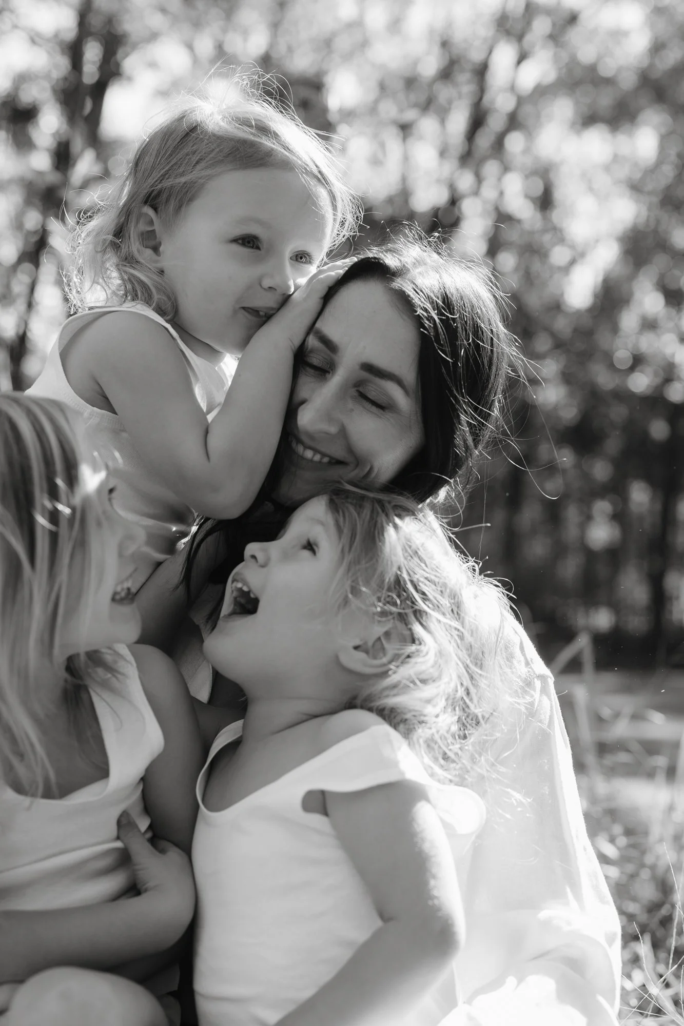 A woman with two young children outdoors, all smiling and laughing, in a joyful moment.