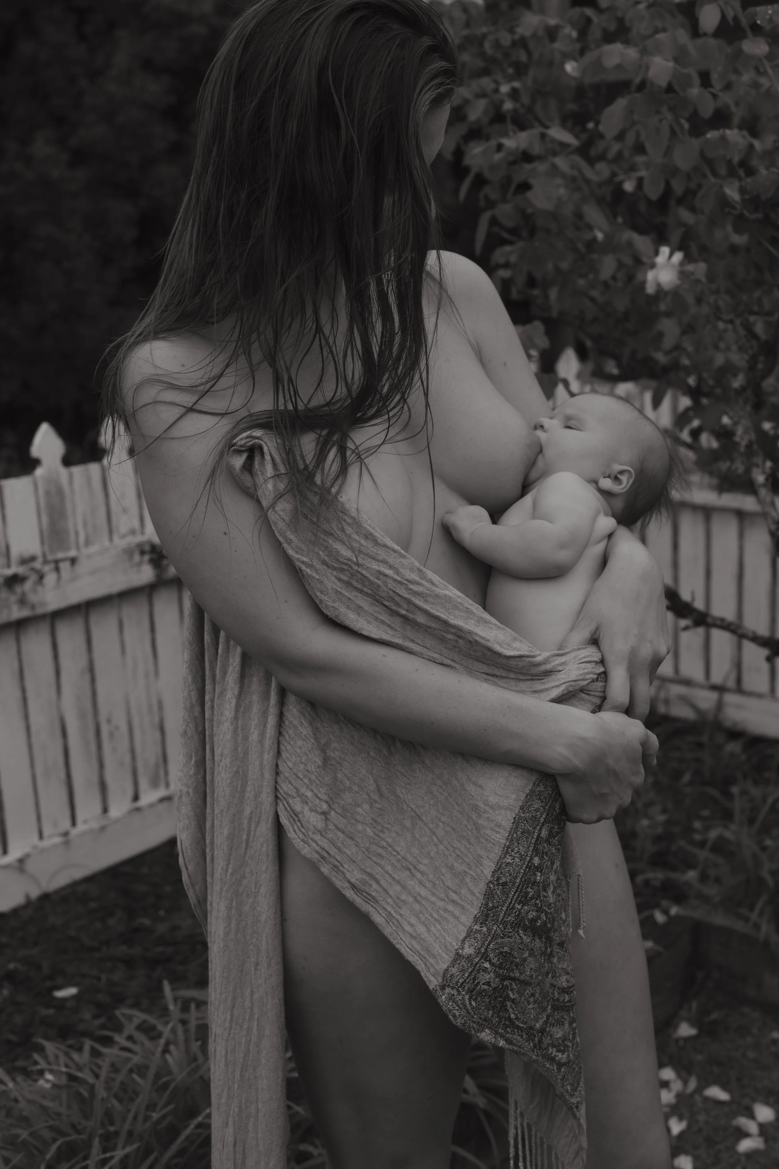 A woman breastfeeding a baby outdoors near a wooden fence and trees, with flowers in the background, in black and white.