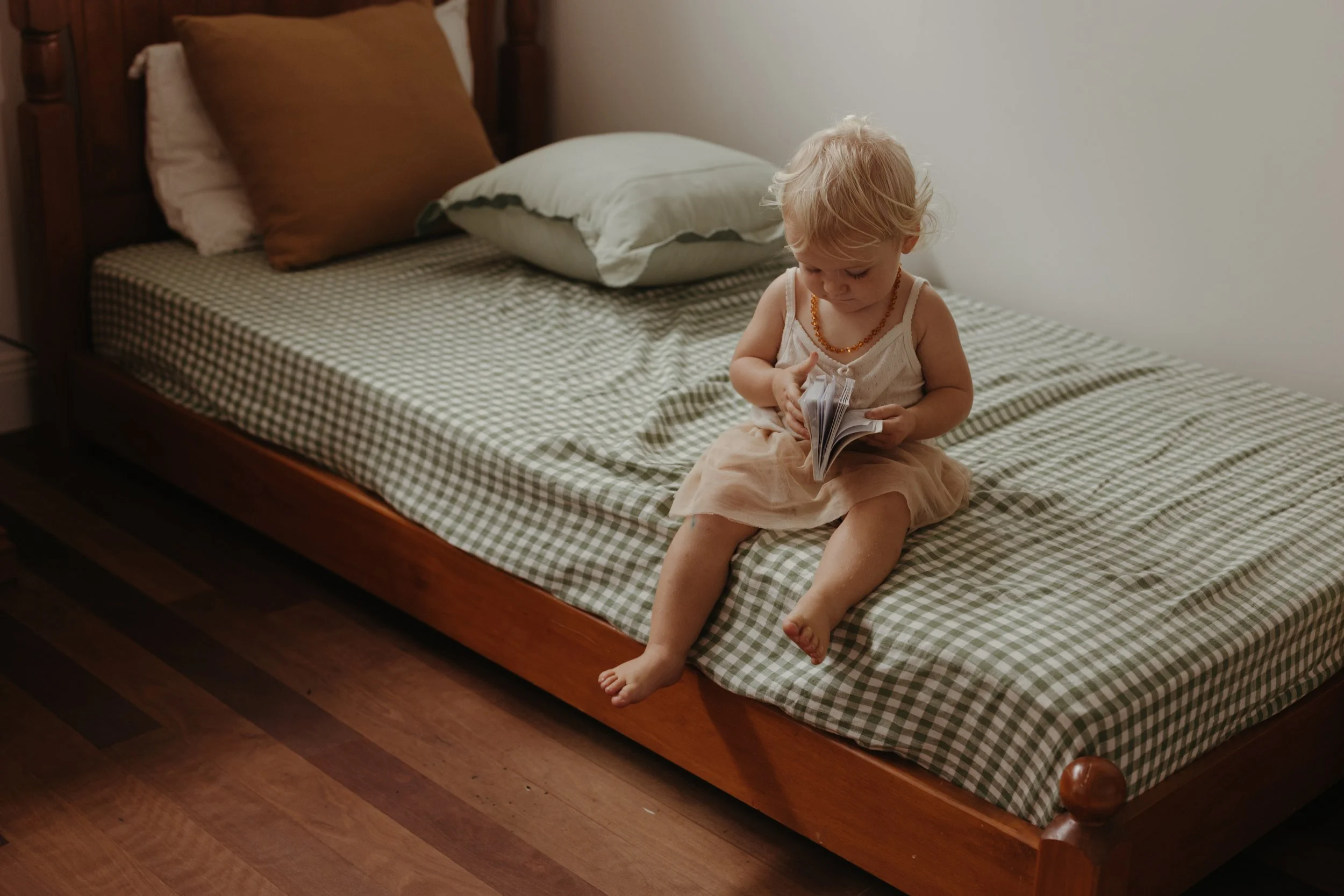 A young child with blonde hair wearing a light-colored dress and a beaded necklace, sitting on a bed and looking at a small book.