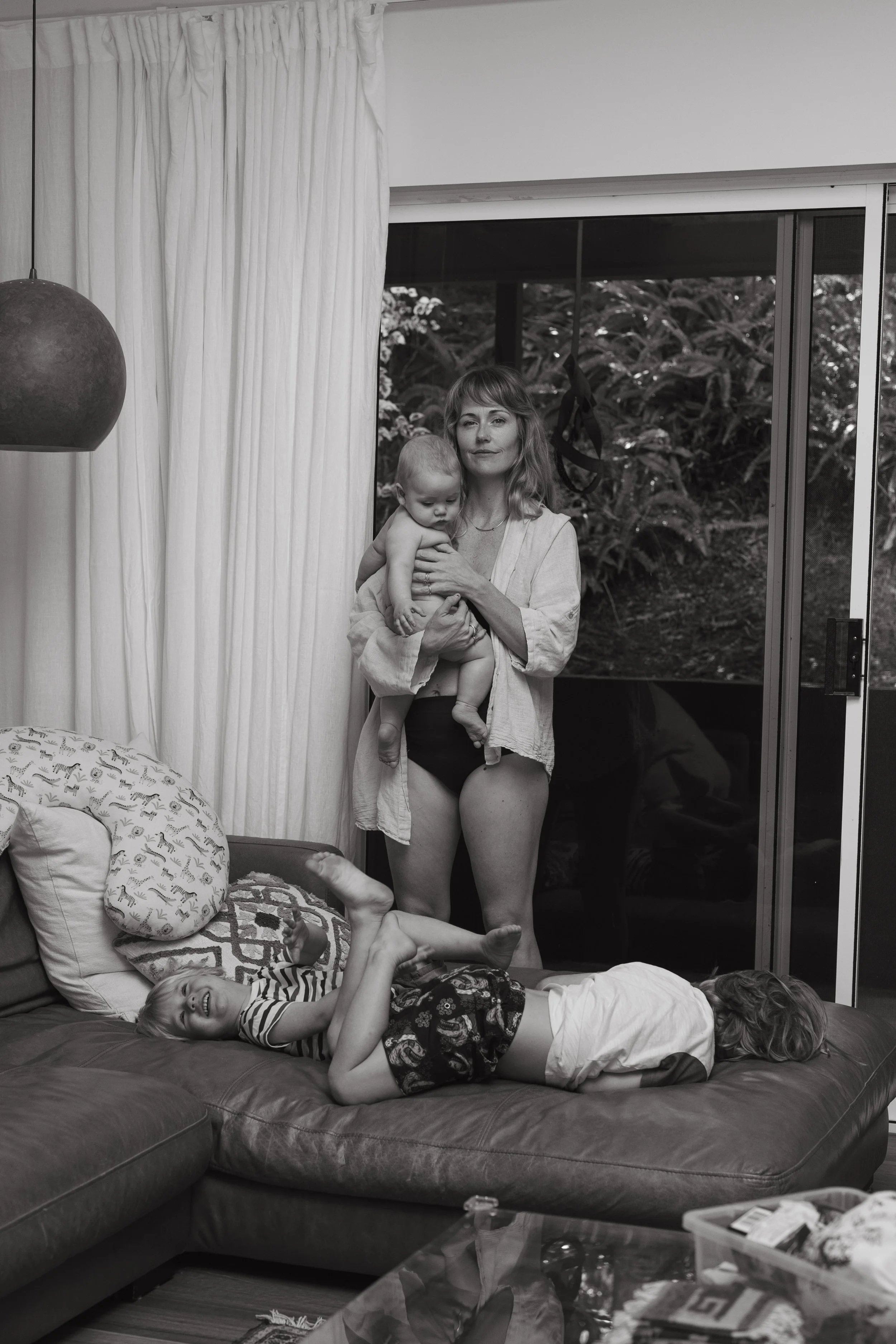 A woman holding a young child standing in a living room, with a child lying on a couch playing, and a sliding glass door with greenery outside.