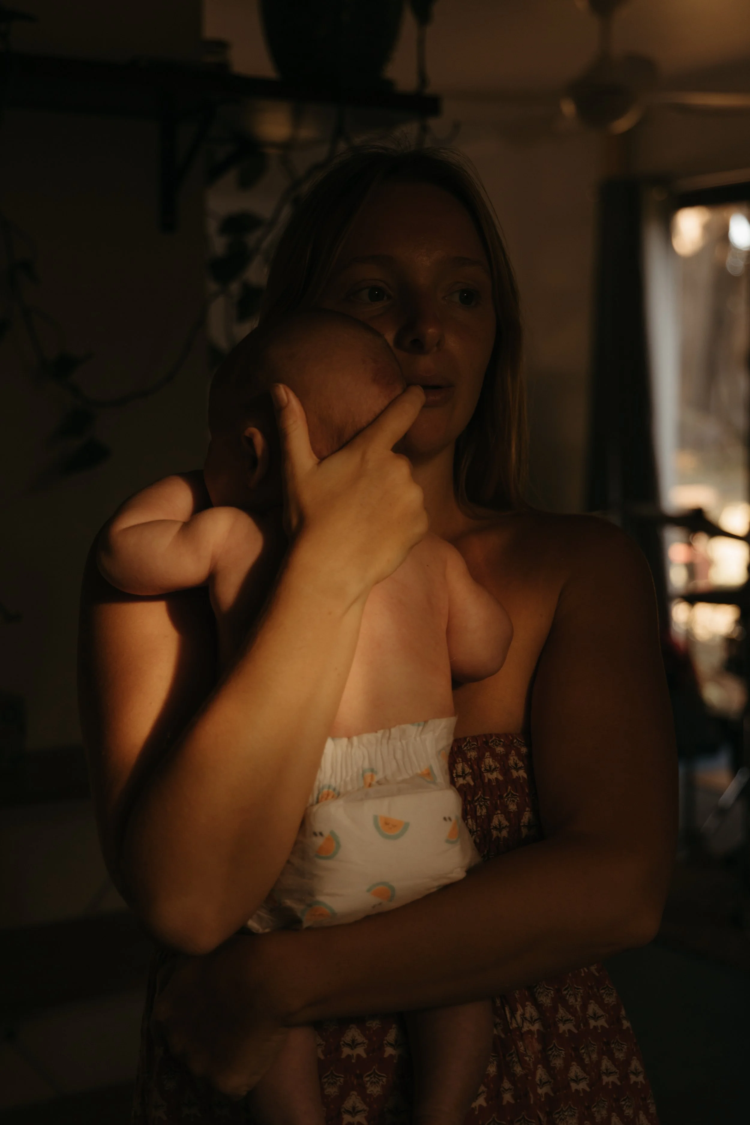 A woman holding a sleeping infant against her chest in a softly lit room.