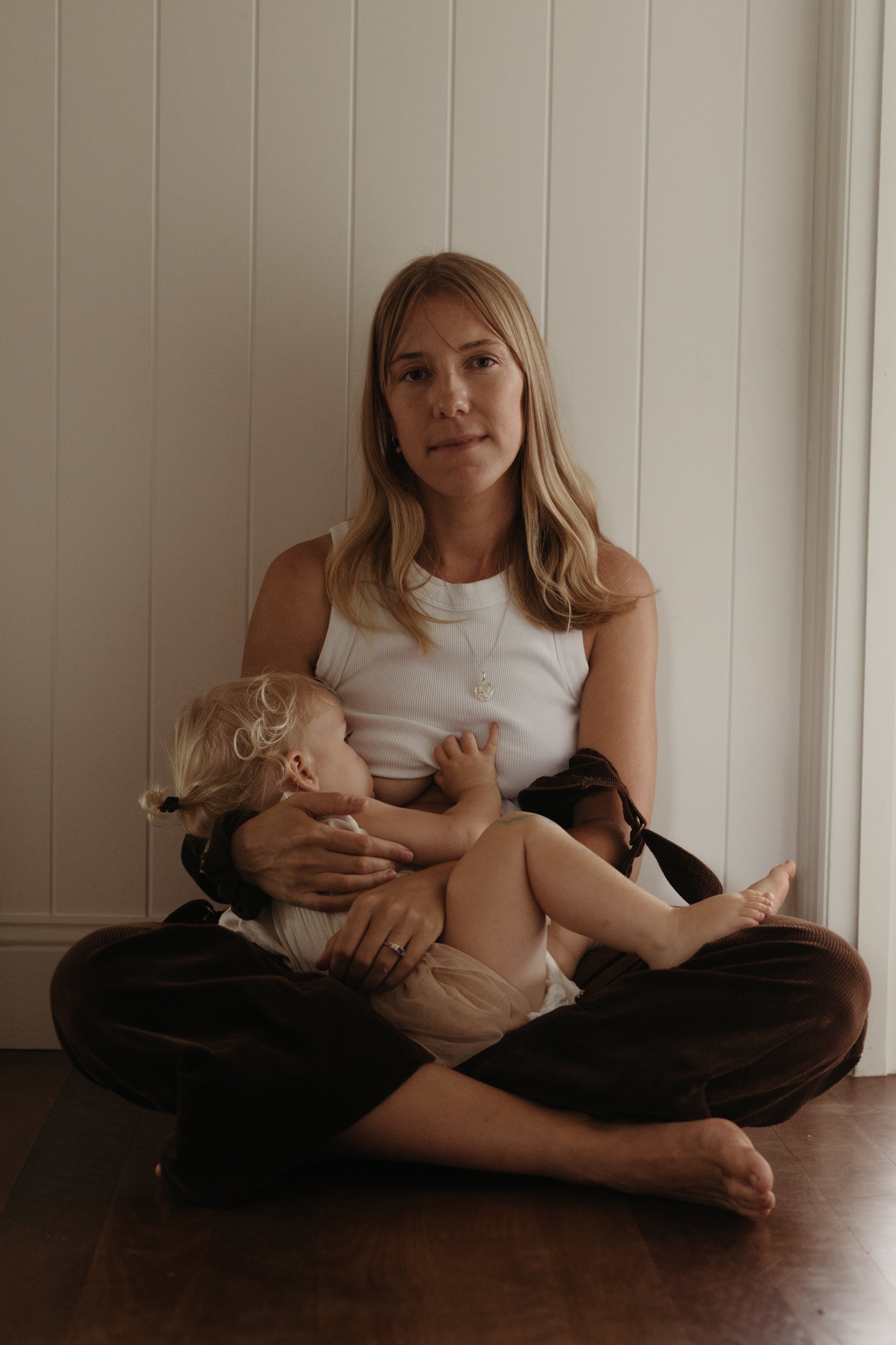 A woman with blonde hair sitting cross-legged on a wooden floor, breastfeeding a young child with blonde curly hair, in front of a white paneled wall.