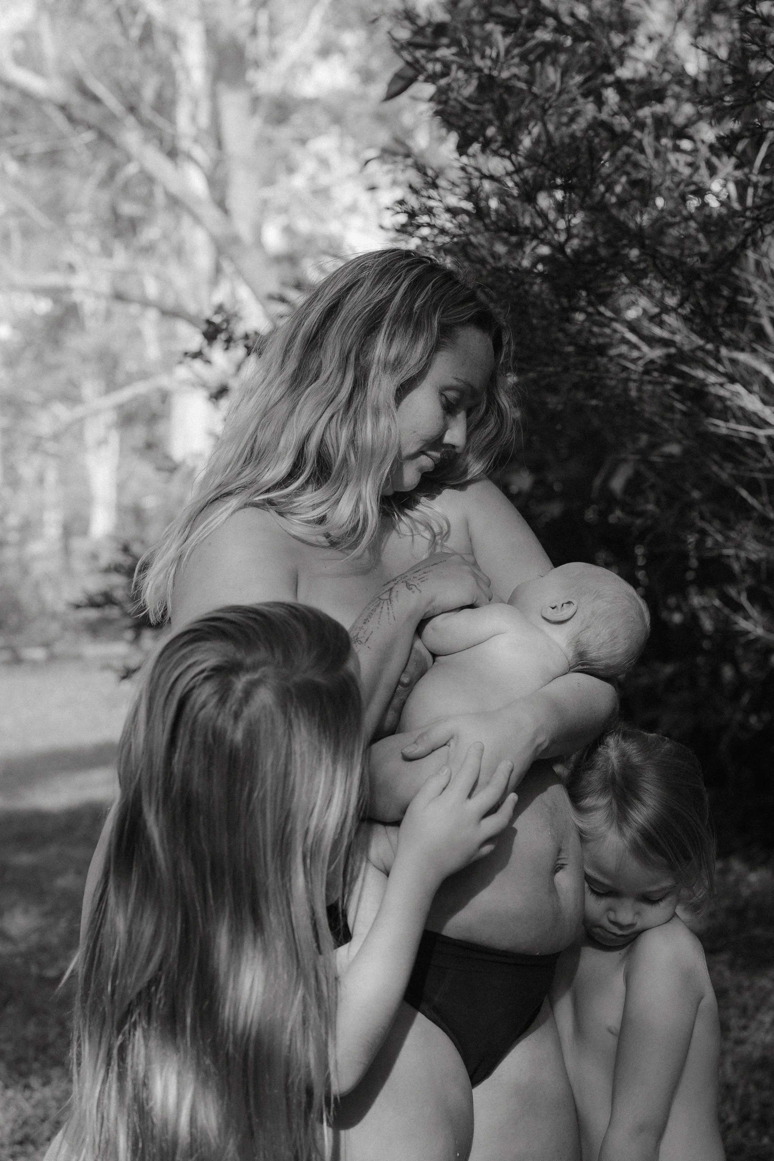 A woman breastfeeding a baby outdoors, surrounded by three young children, in a natural setting.