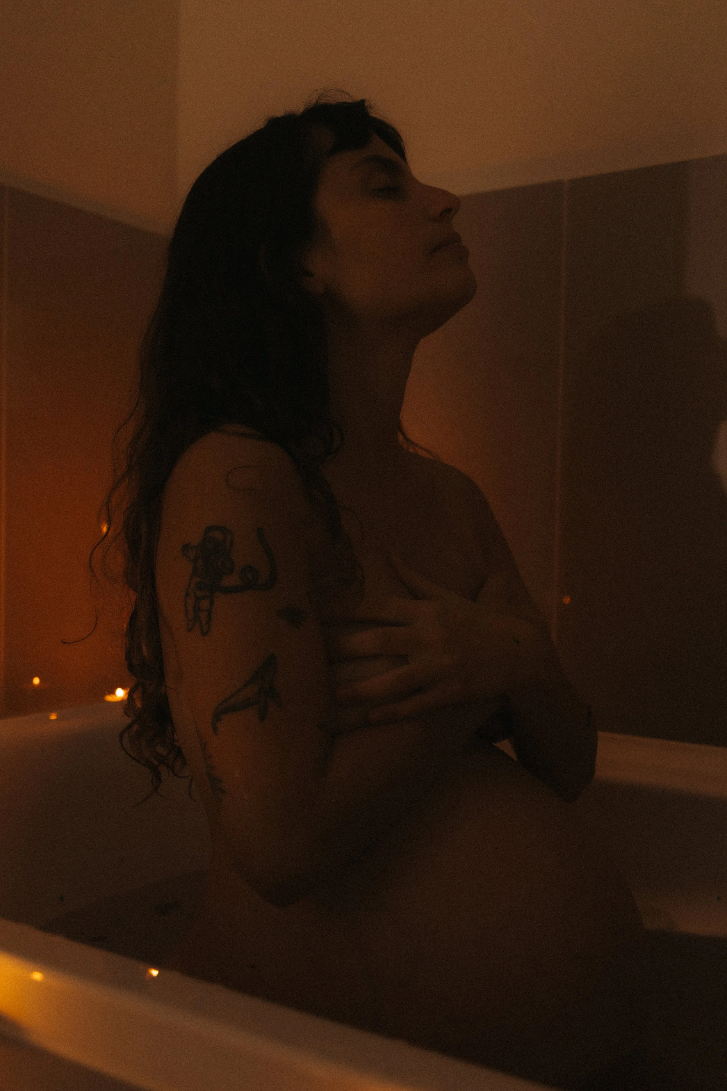 A woman with tattoos on her arm sitting in a dimly lit bathtub with her eyes closed, holding her chest, in a warm-toned room.