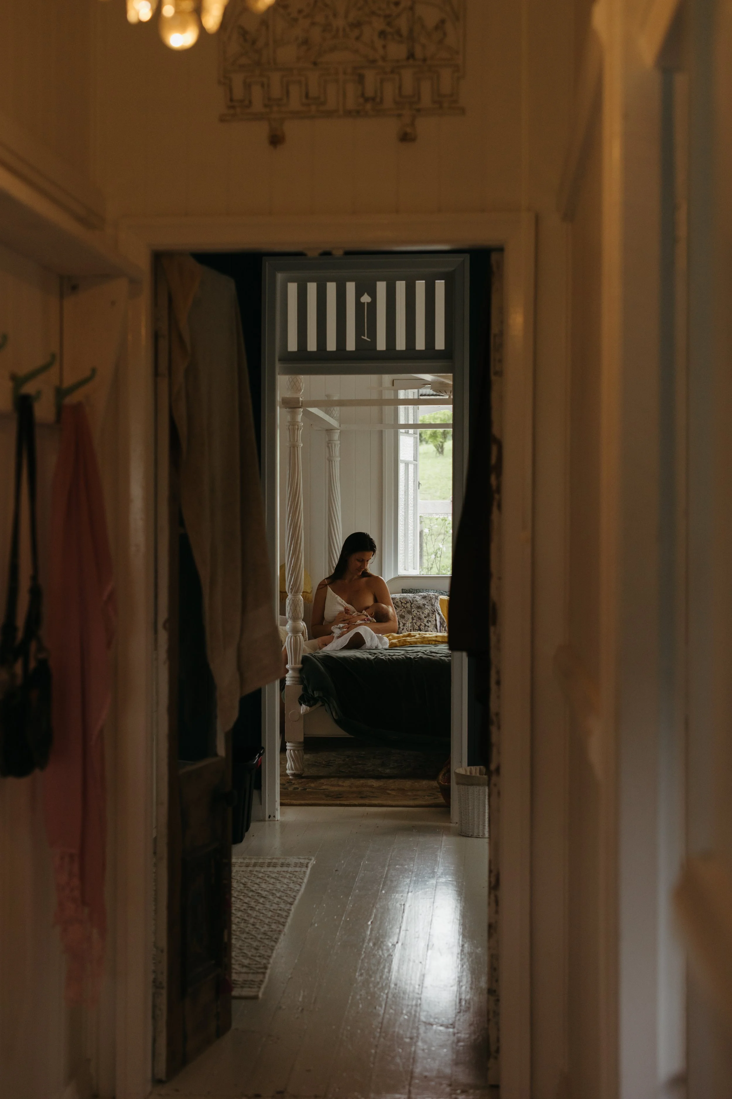 View through a doorway into a bedroom with a woman sitting on a bed by a large window, nursing a baby, with natural light coming in.