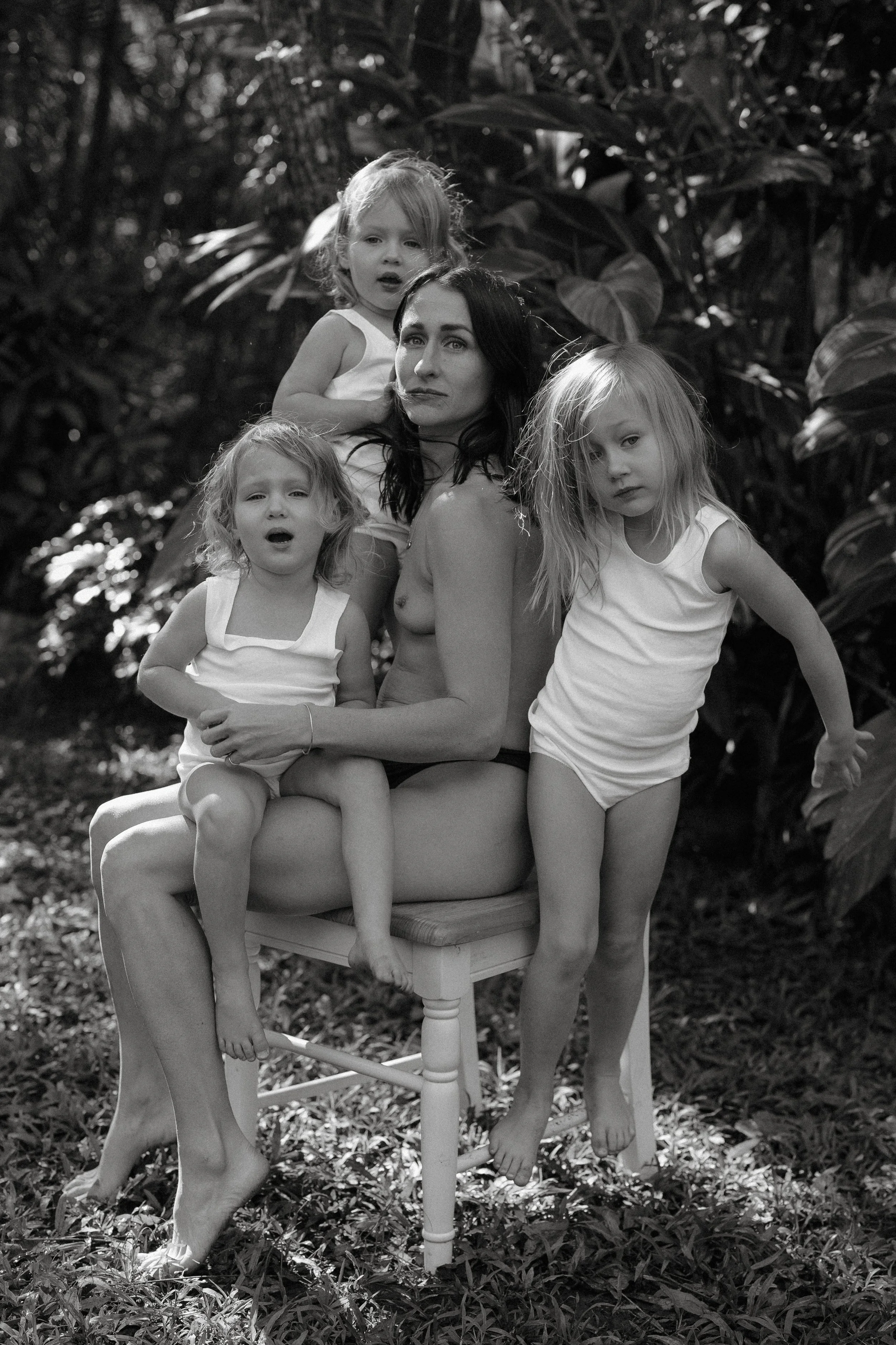 Black and white photo of a woman surrounded by four young girls, outdoors with large leafy plants in the background. The woman is sitting on a chair, with three of the girls on her lap and one standing beside her, all wearing white tank tops or dress