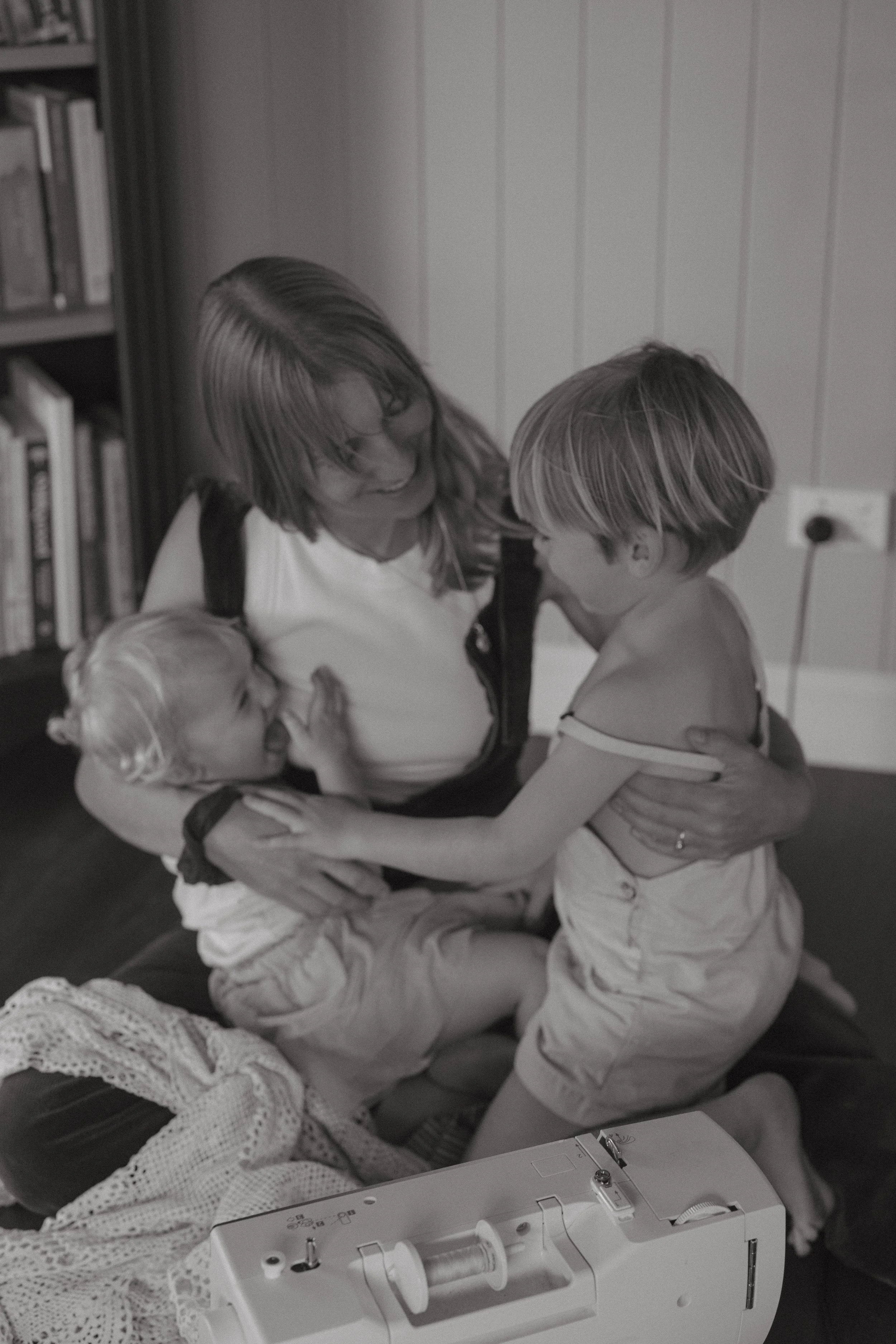 A woman with two young children on a bed, with one child sitting and the other standing, engaging in a playful moment.