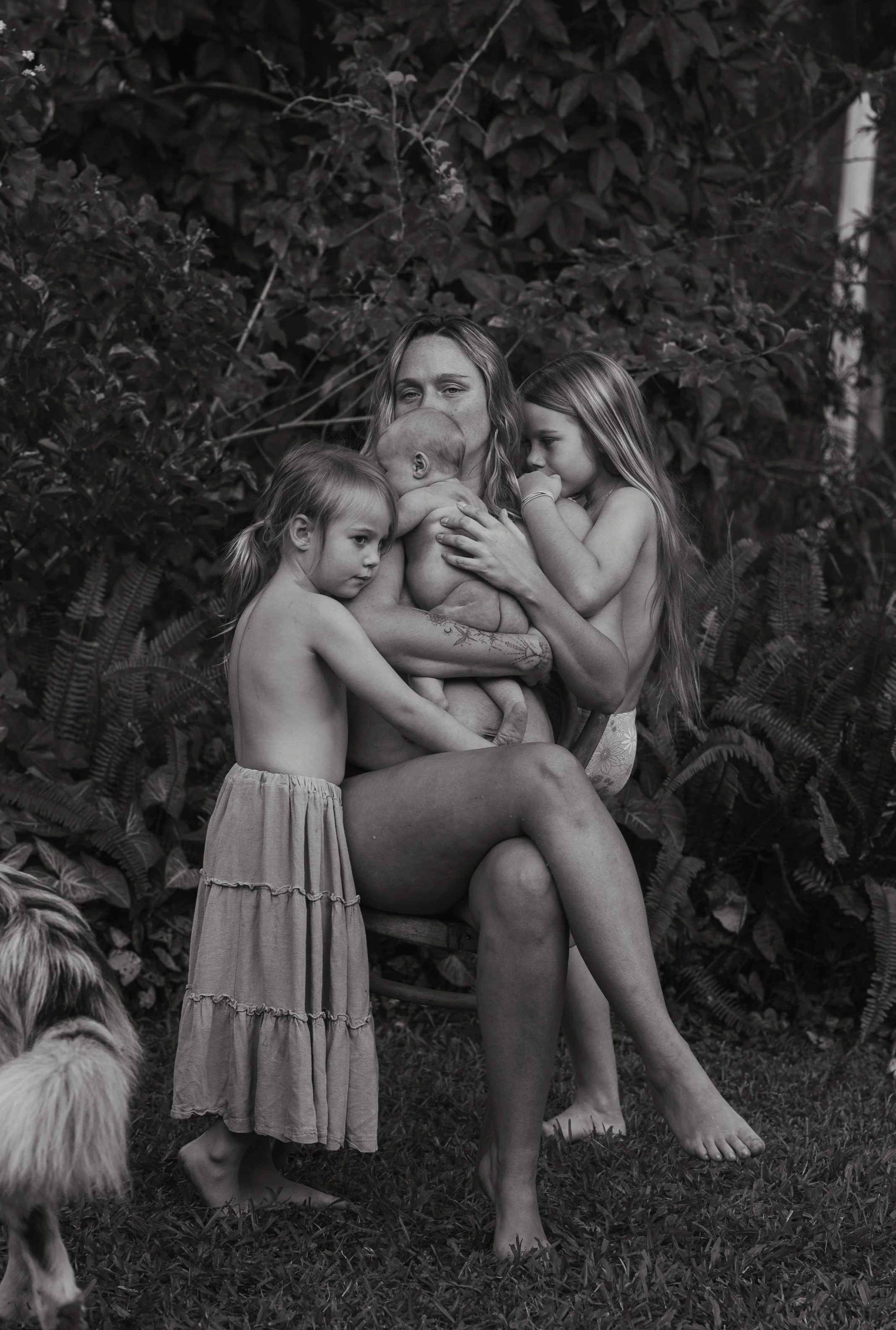 A woman surrounded by three young girls and a dog outdoors near lush foliage. The woman is holding a baby and others are close to her, creating an intimate scene.