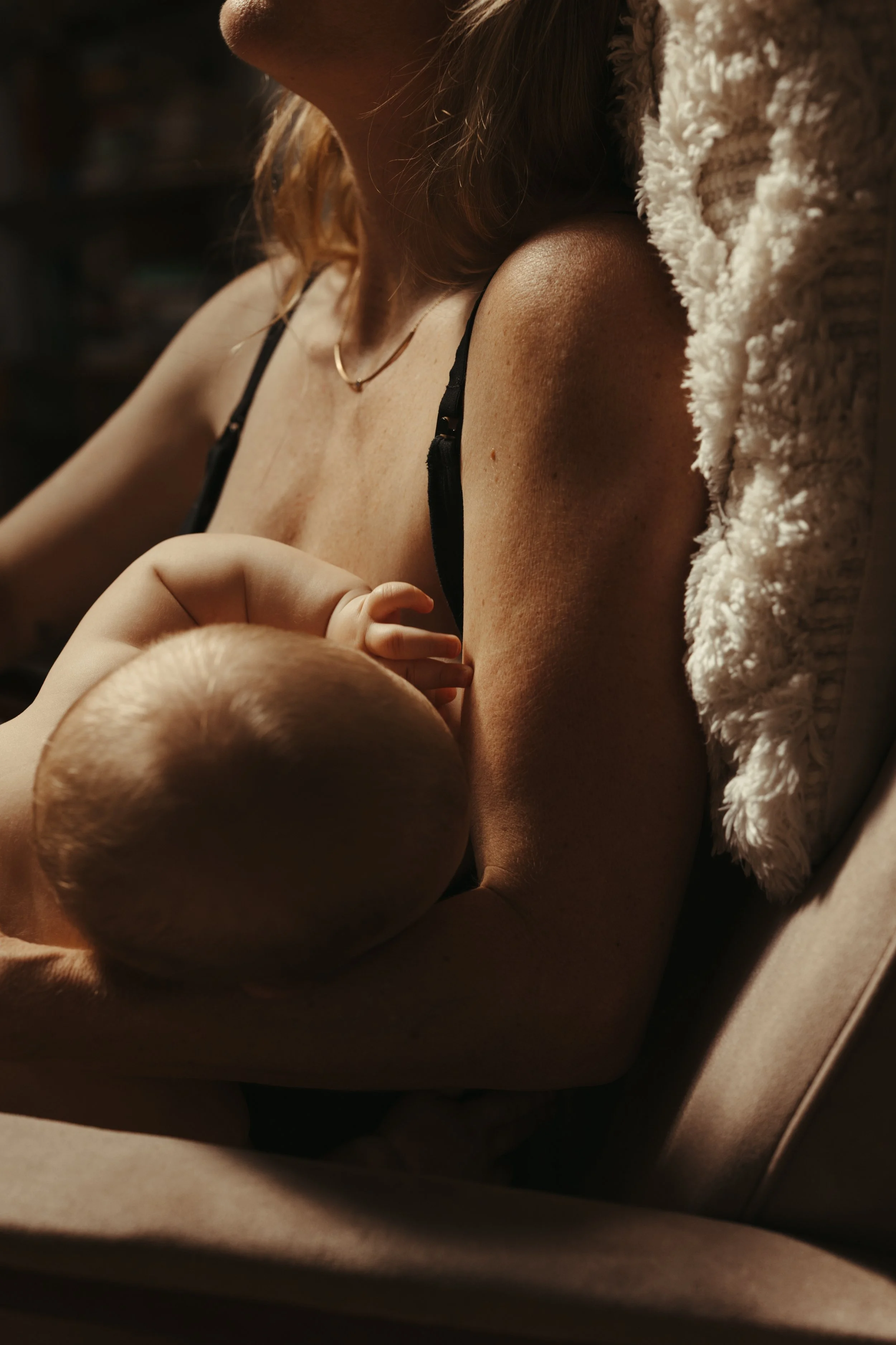 A woman breastfeeding a baby in a cozy setting, close-up shot showing the woman's shoulder, arm, and part of her face, with the baby nursing in her embrace.