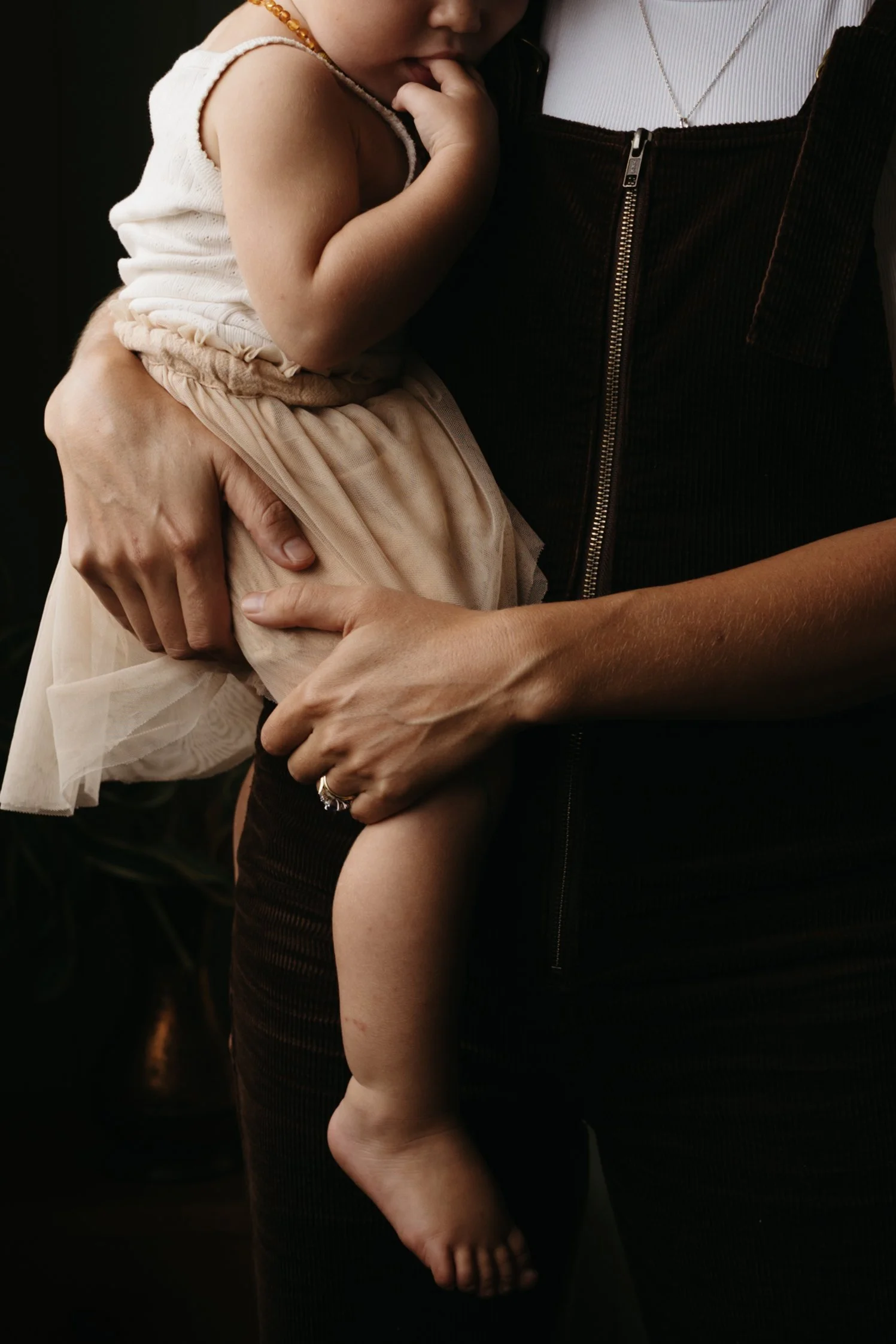 A person is holding a young child, with the child resting their head on the adult's shoulder. The adult's hands gently support the child's waist and leg, and the child's arm is around the adult's neck, with fingers near their mouth.