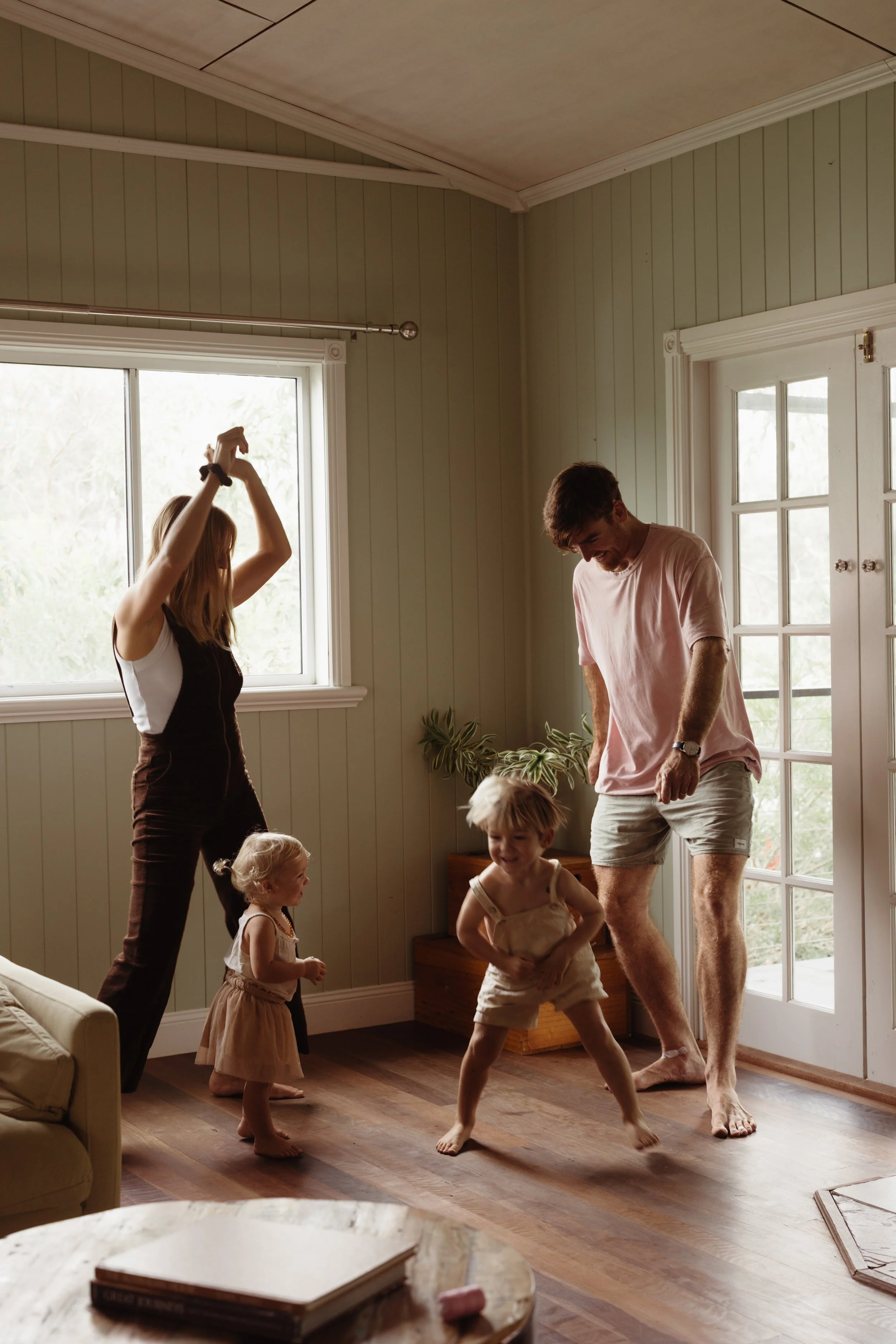 A family of four, including two young children, is dancing joyfully inside a living room near a large window and a glass door with sunlight streaming in.