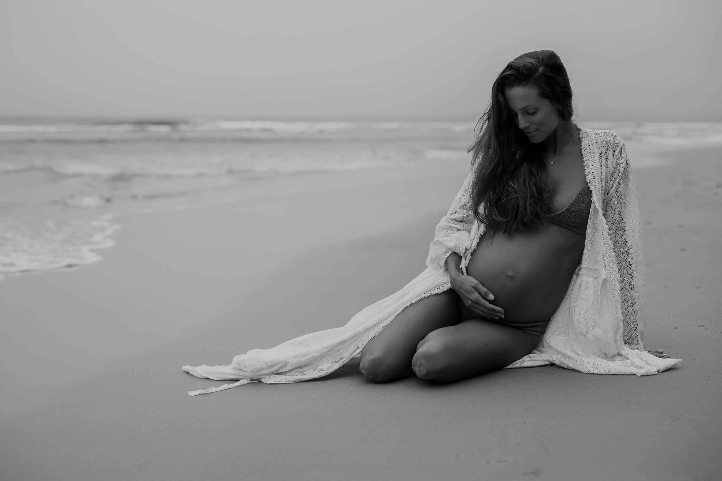 A pregnant woman kneeling on the beach, touching her belly, wearing a bikini and a long lace cover-up, with the ocean in the background.