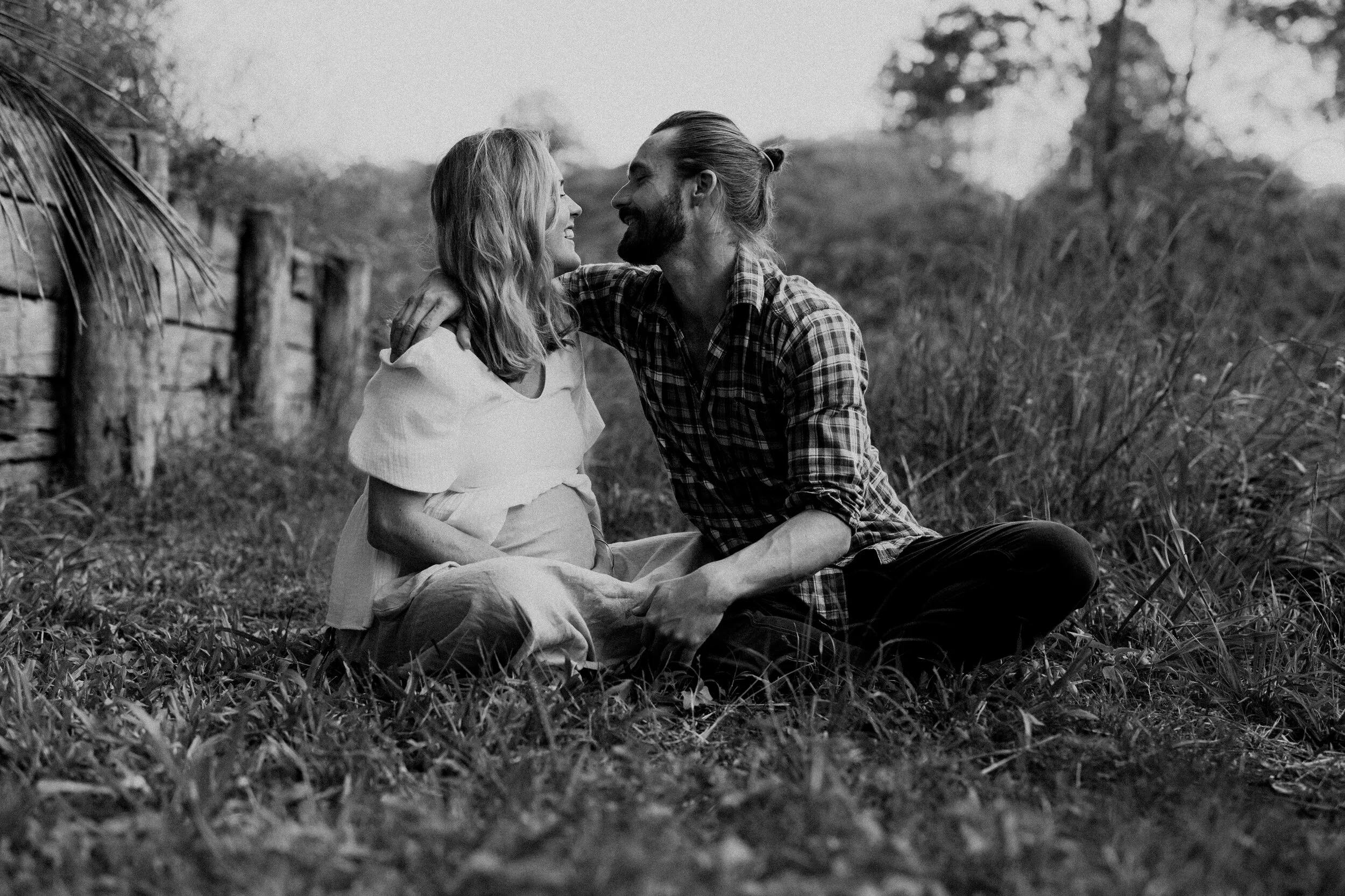 A black and white photo of a couple sitting on the grass, smiling and looking at each other, with an old wooden fence and trees in the background.