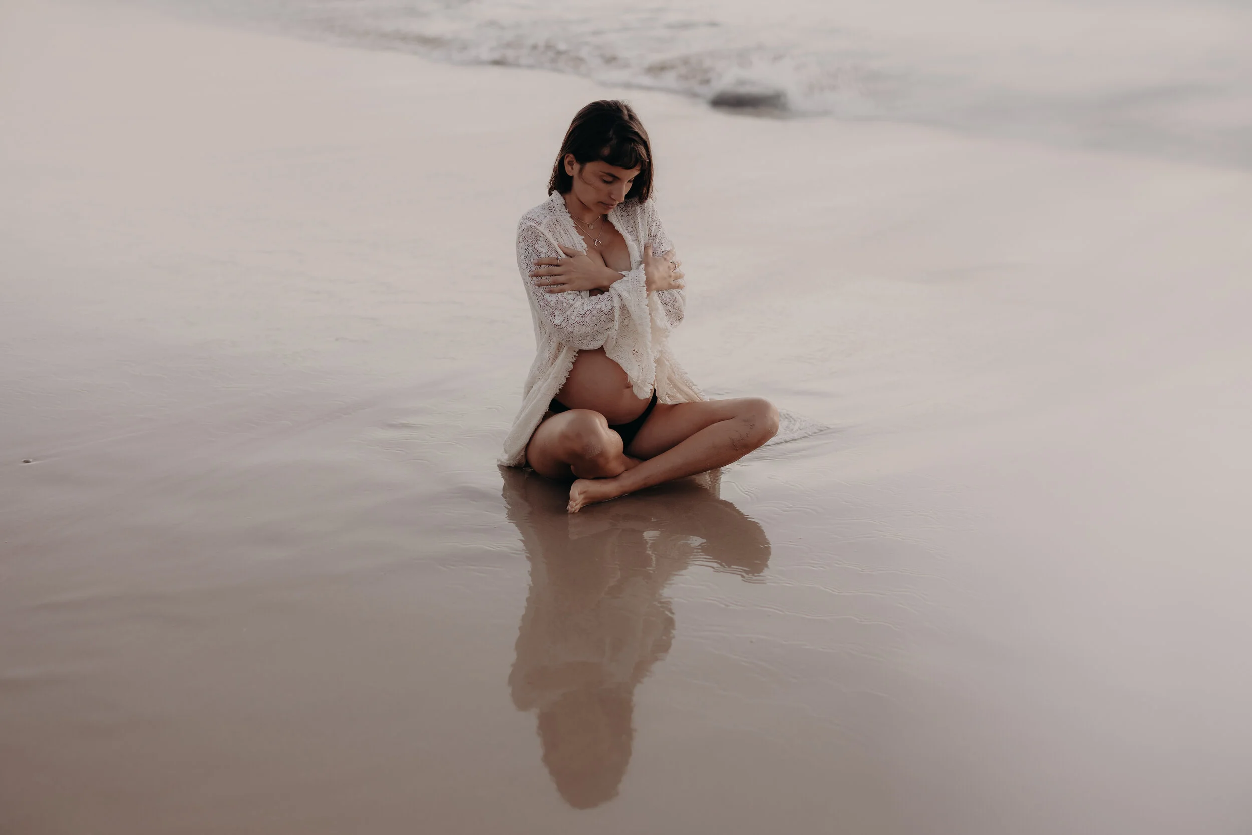 A pregnant woman sitting on the wet sand at the beach, cradling her belly and looking down, wearing a white lace cover-up with a black bikini underneath. The reflection of her is visible in the shallow water.