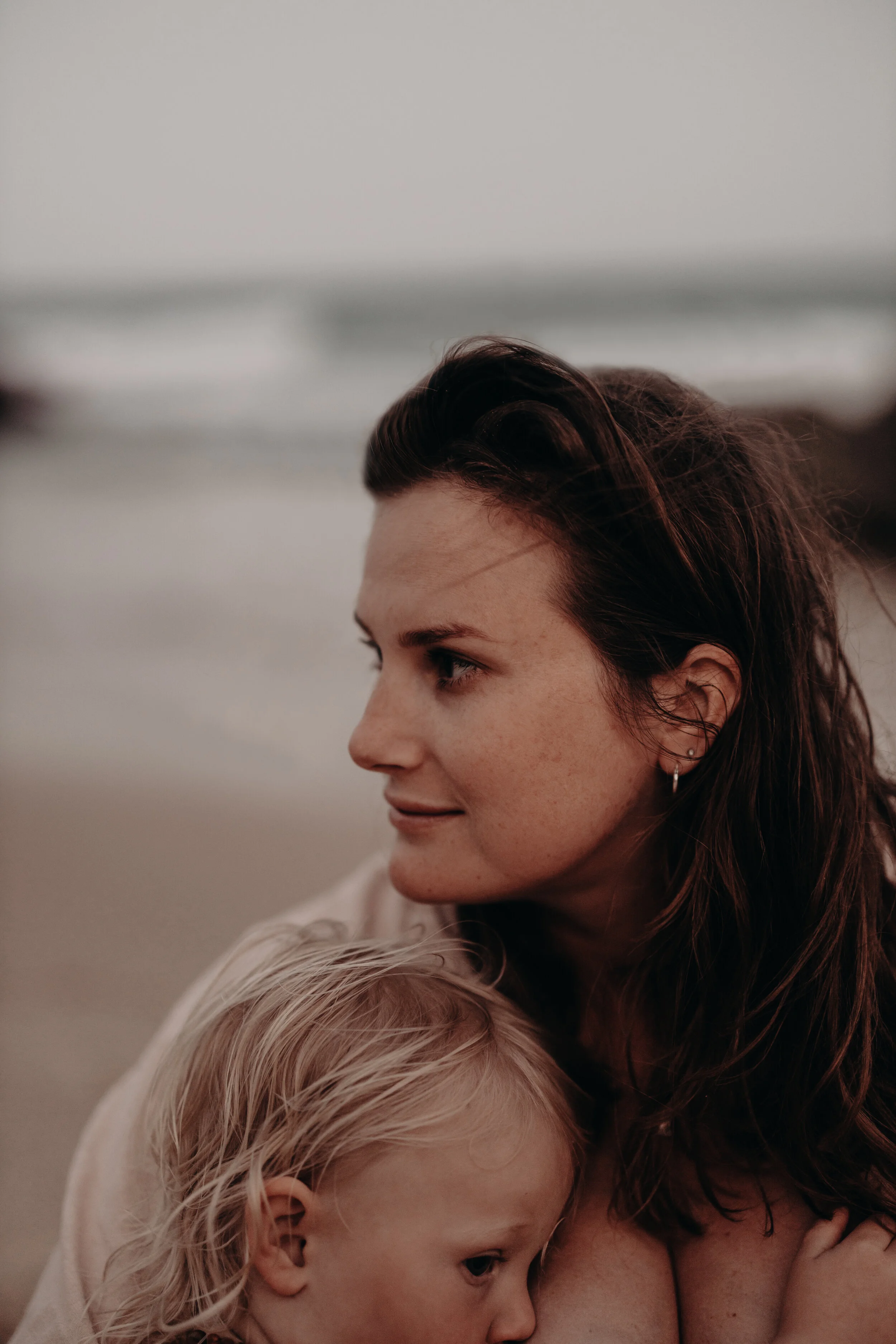 A woman with brown hair and a young blonde girl close together on a beach, with the ocean visible in the background.