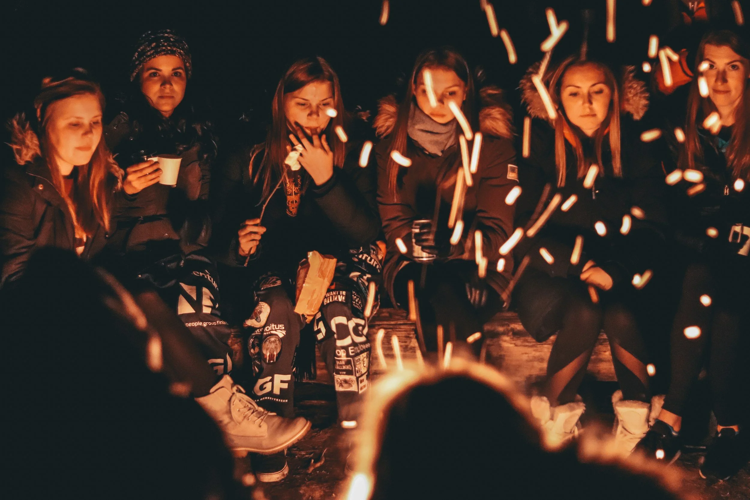 Group of young women sitting around a campfire at night, some holding mugs, with sparks flying from the fire.