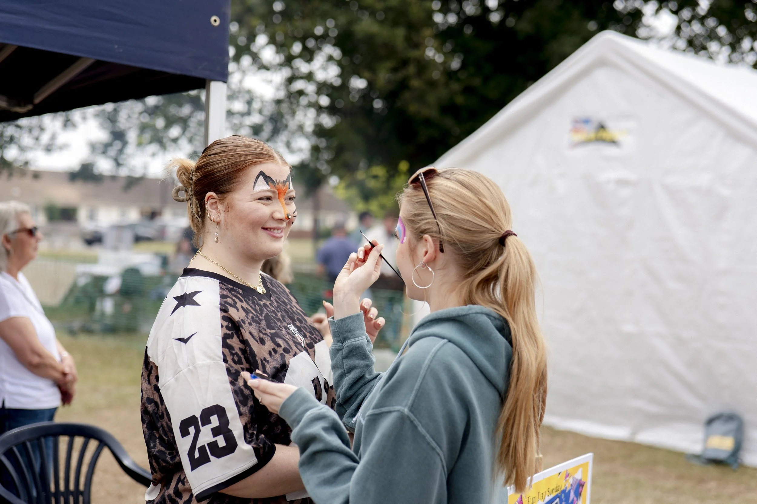 A woman with face paint of a fox is being painted by another woman at an outdoor event, with tents and people in the background.