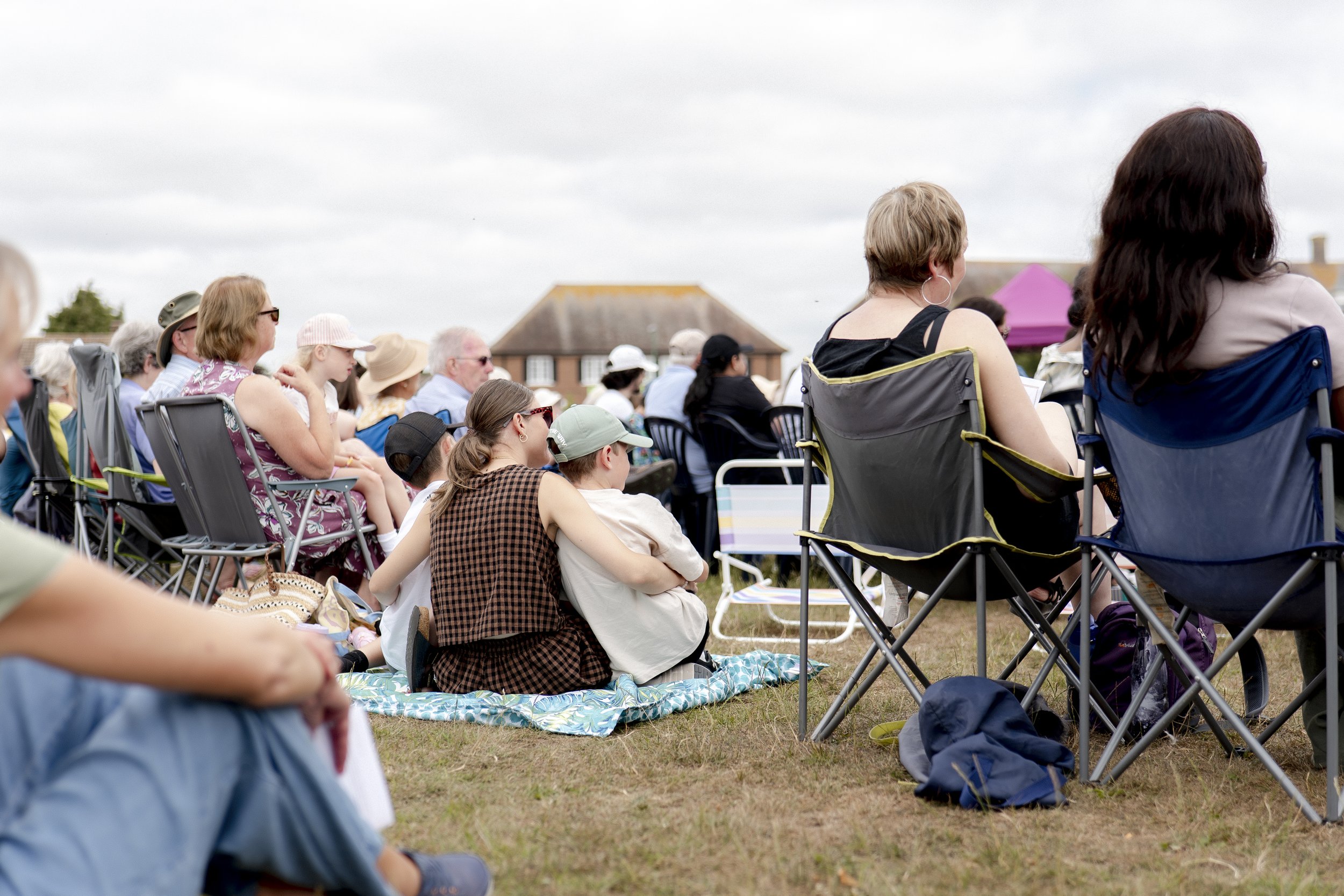 A group of people sitting outdoors on a grassy field, watching an event. They are seated on lawn chairs and blankets, with some wearing hats and sunglasses, under a partly cloudy sky.