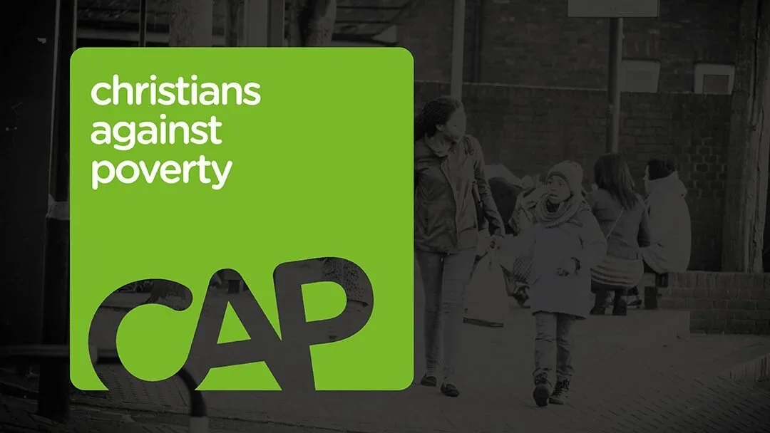A green sign with white and black text that reads 'Christians against poverty' and includes the abbreviation 'CAP' with the 'A' stylized as a triangle. In the background, there are people sitting and standing outside a building.
