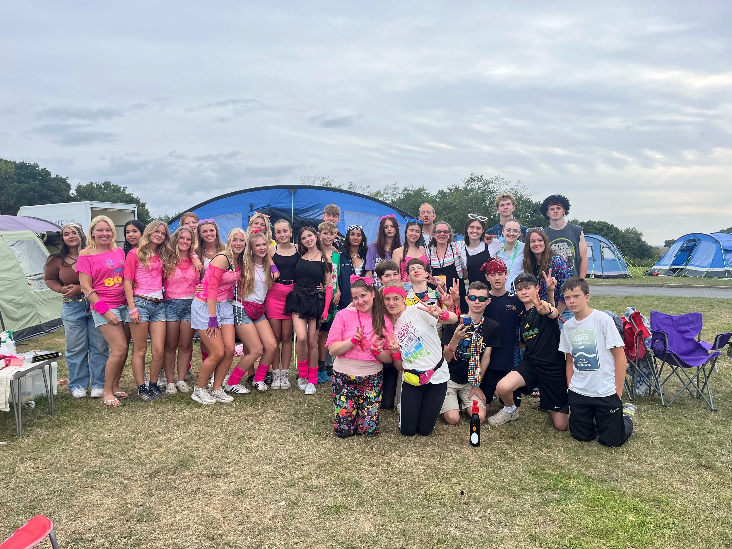 Group of teenagers and young adults at a campsite with tents in the background, dressed in colorful and casual clothing, posing for a group photo outdoors on a cloudy day.