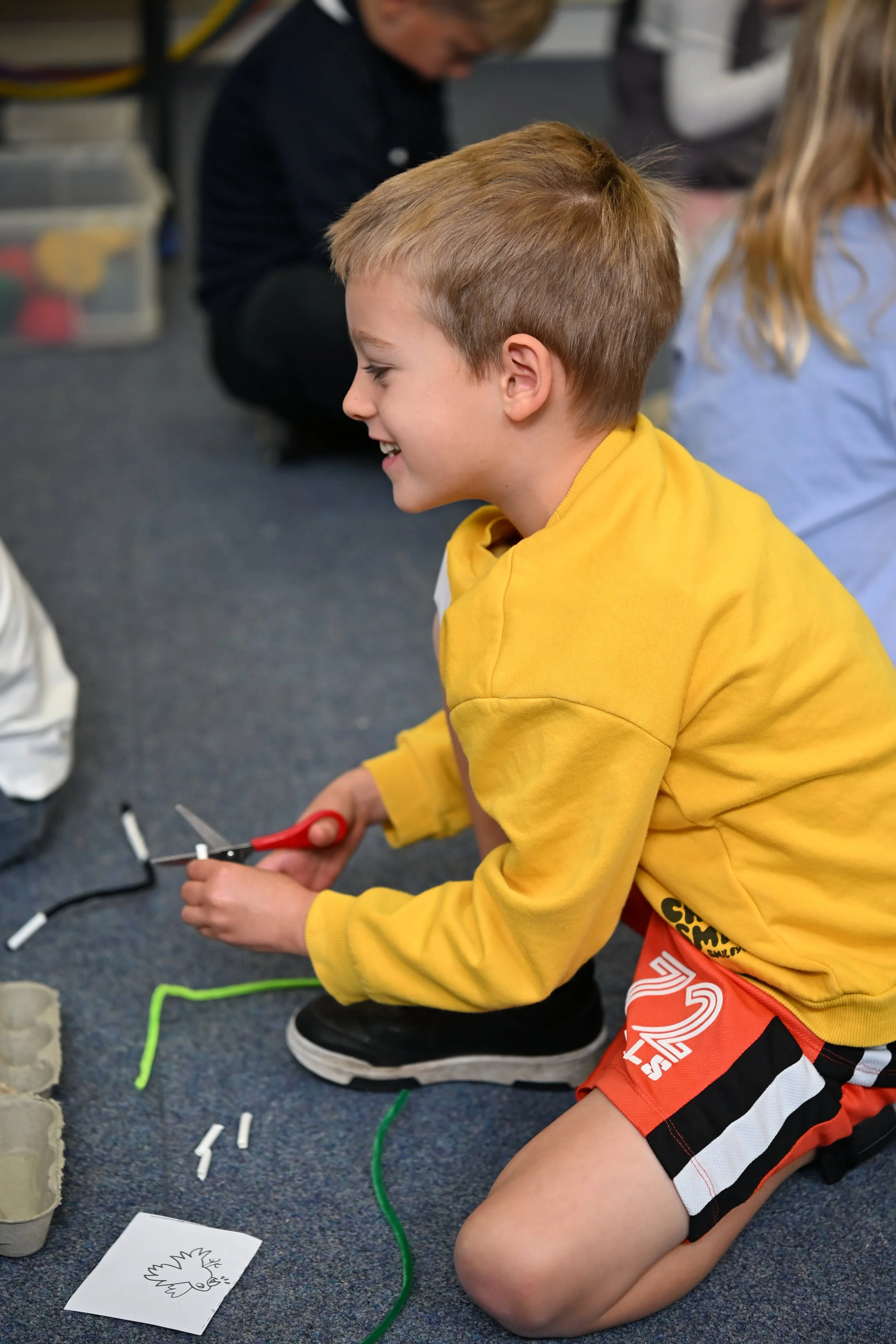A young boy with blonde hair wearing a yellow shirt and orange shorts kneeling on a classroom carpet, using scissors on a craft project, surrounded by craft materials and wire, with other children in the background.