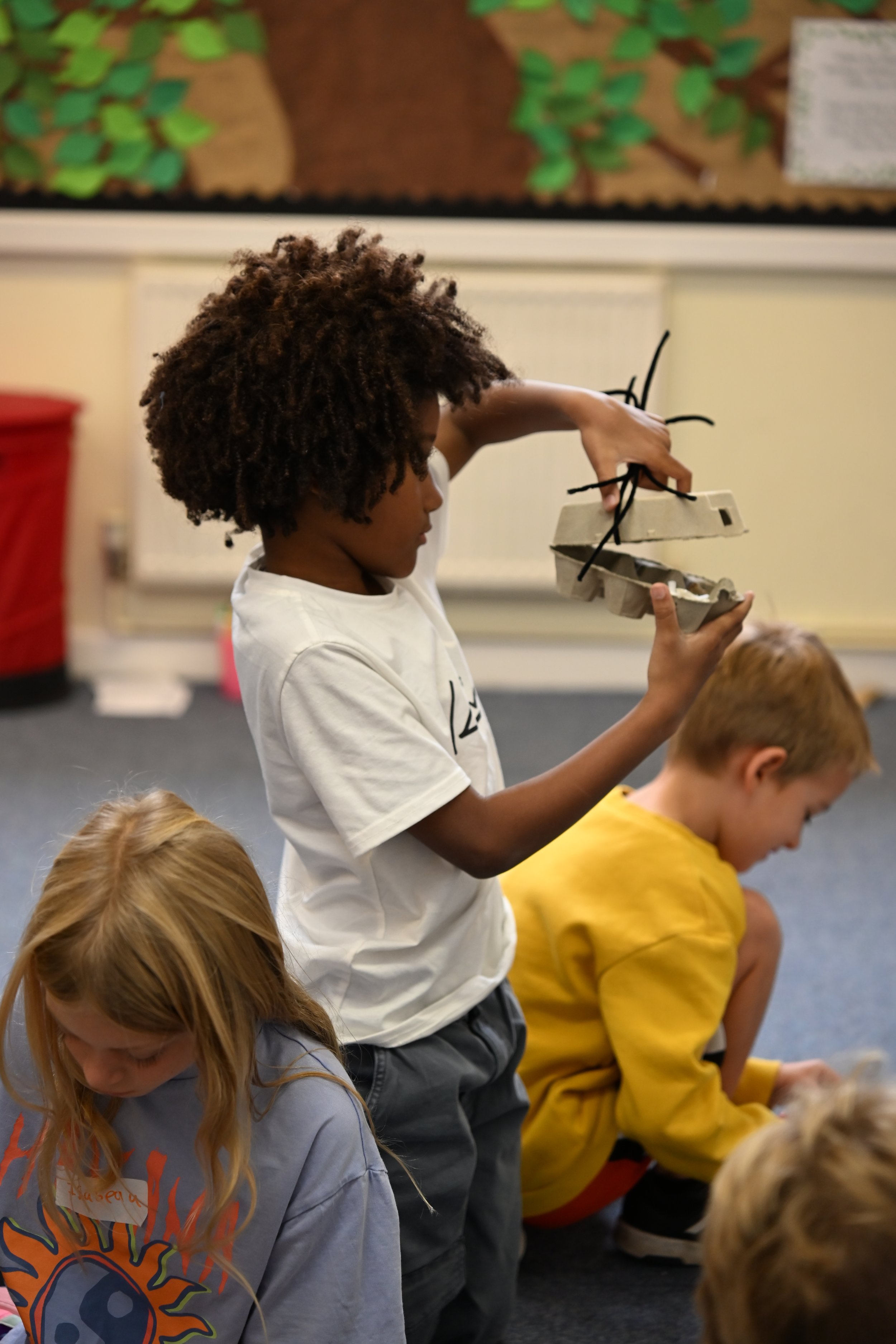 A young boy with curly hair standing and holding a cardboard and black sticks, possibly a craft project, while other children sit on the floor in front of him in a classroom.