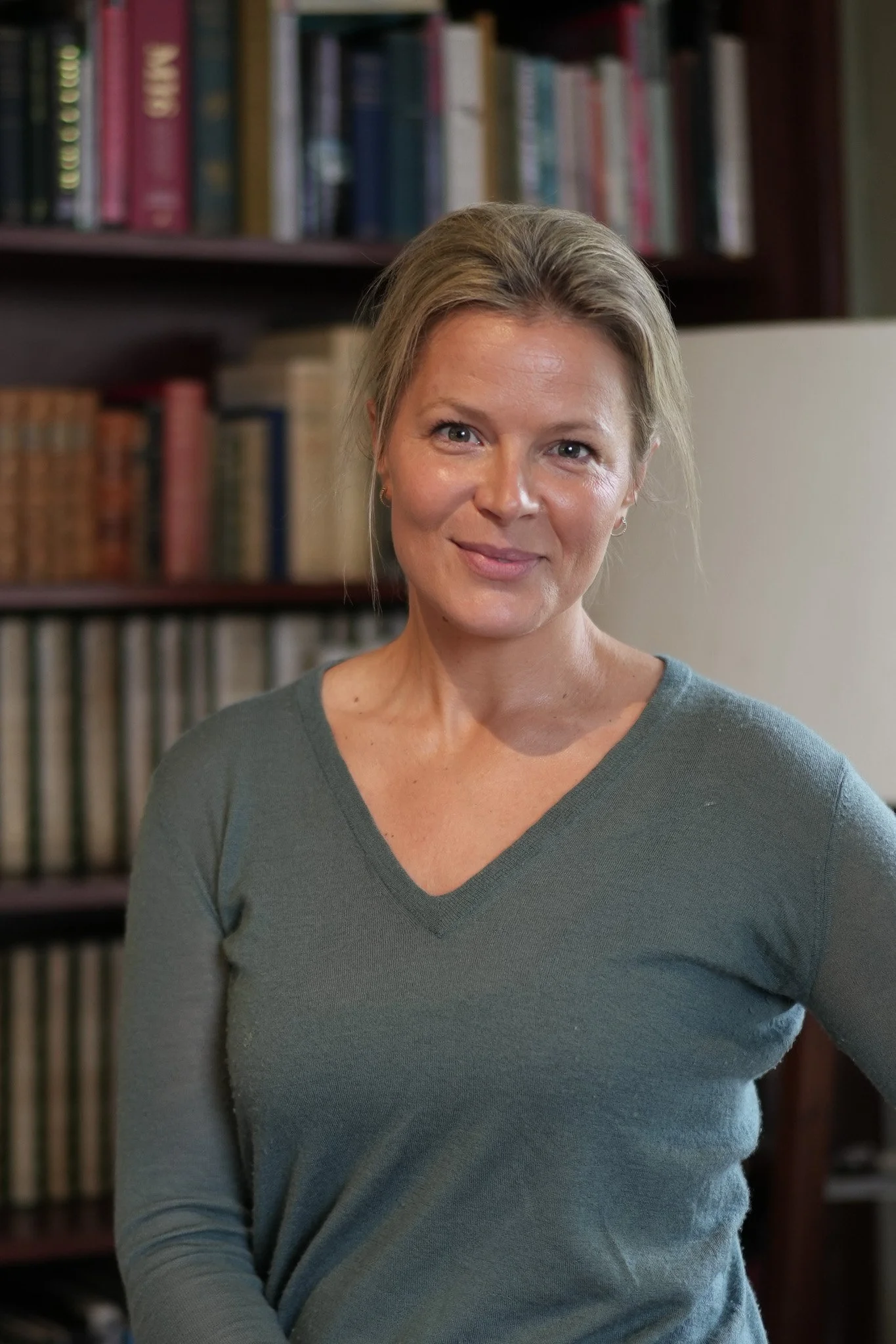 A woman with blonde hair wearing a gray V-neck sweater standing in front of a bookshelf filled with books.